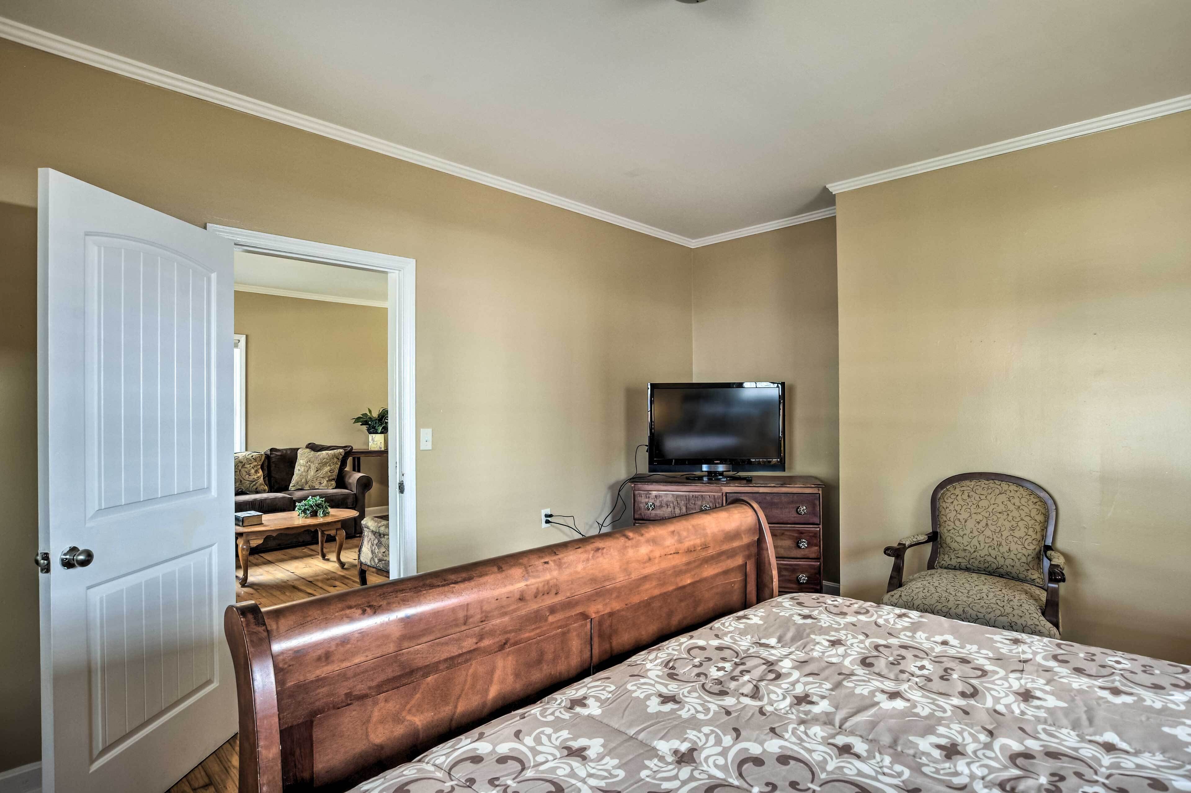A bedroom with a wooden headboard bed, a television on a dresser, a chair, and a door leading to another room.