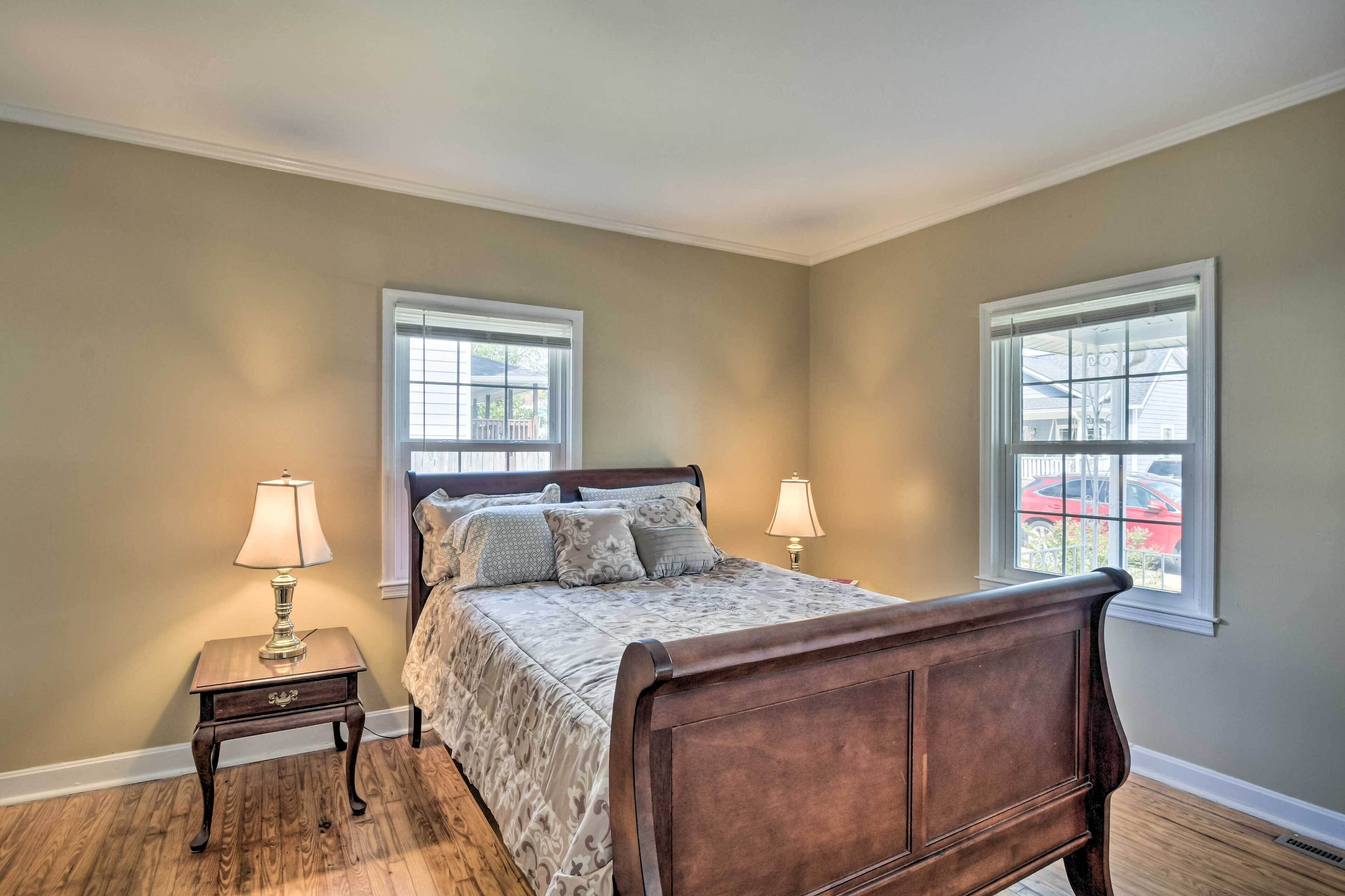 A bedroom with a wooden bed, two bedside lamps, a nightstand, and two windows with blinds.