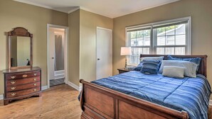 A bedroom with a wooden bed, a dresser, a mirror, a window with blinds, and a door leading to a bathroom.