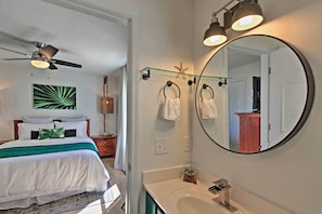 A bedroom with a bed, a ceiling fan, a green wall art, a nightstand, and a bathroom with a sink, mirror, and towel rings. - Emerald Hideaway Renovated Condo in Navarre Beach (Navarre)