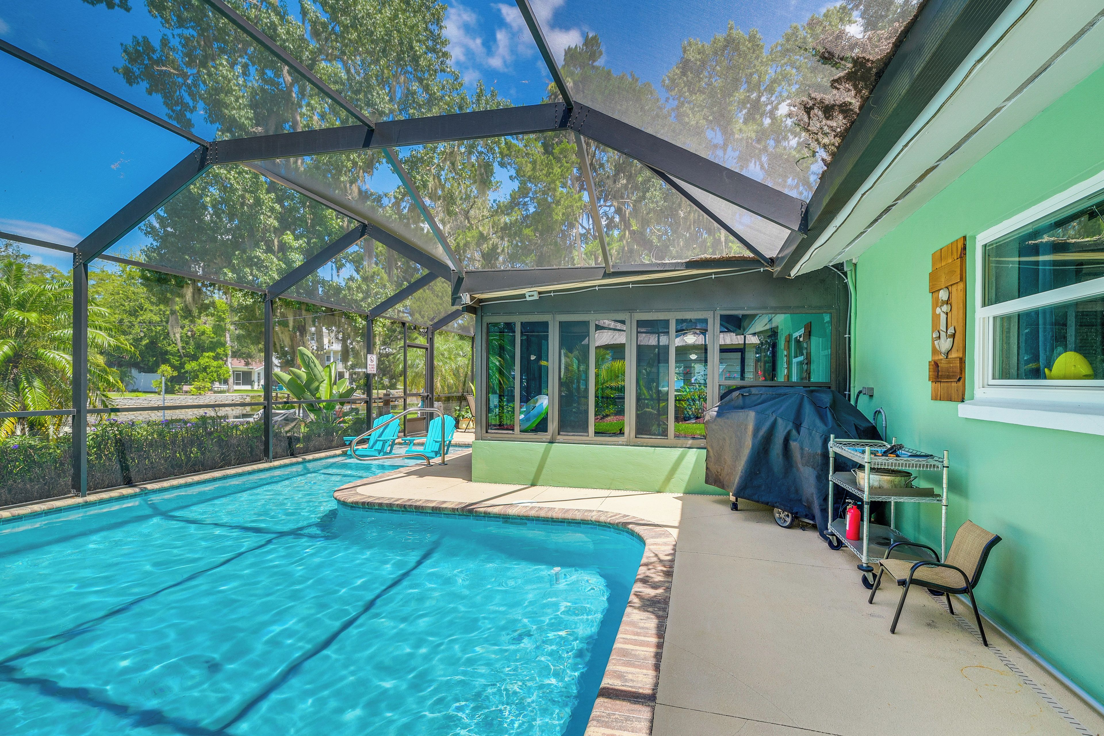 A covered outdoor swimming pool with clear blue water, surrounded by a metal frame and glass panels, and a patio area with a table and chairs.