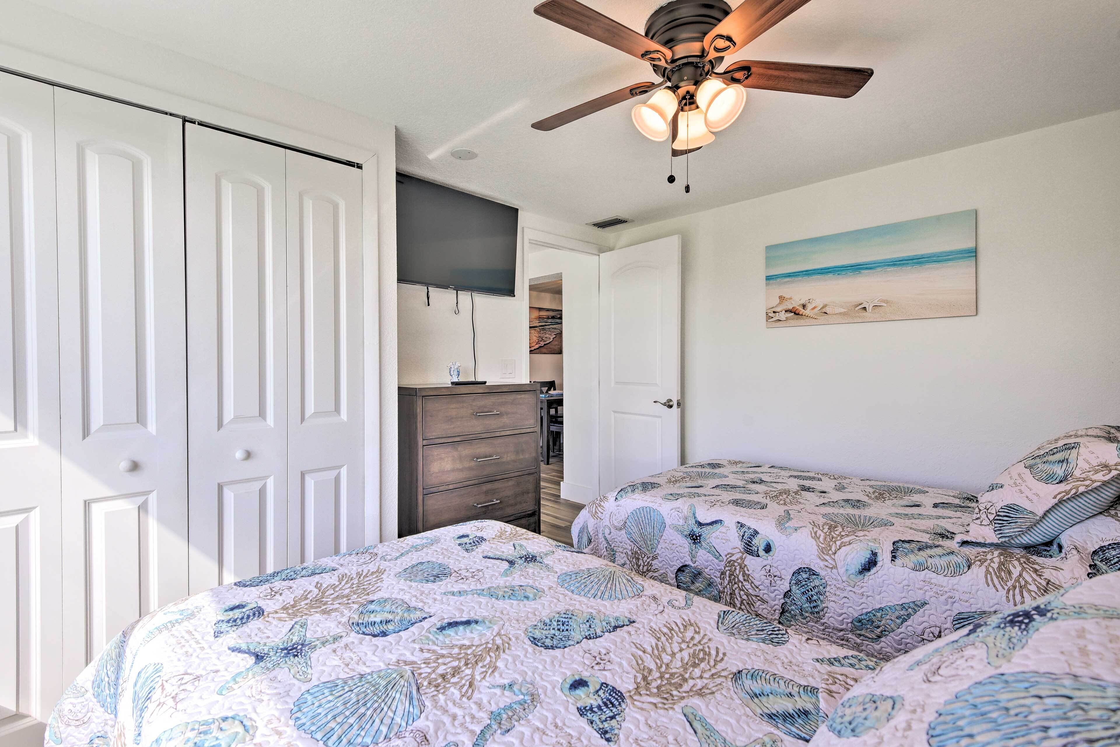 A bedroom with two beds, a ceiling fan, a painting of a beach scene, and a wardrobe.