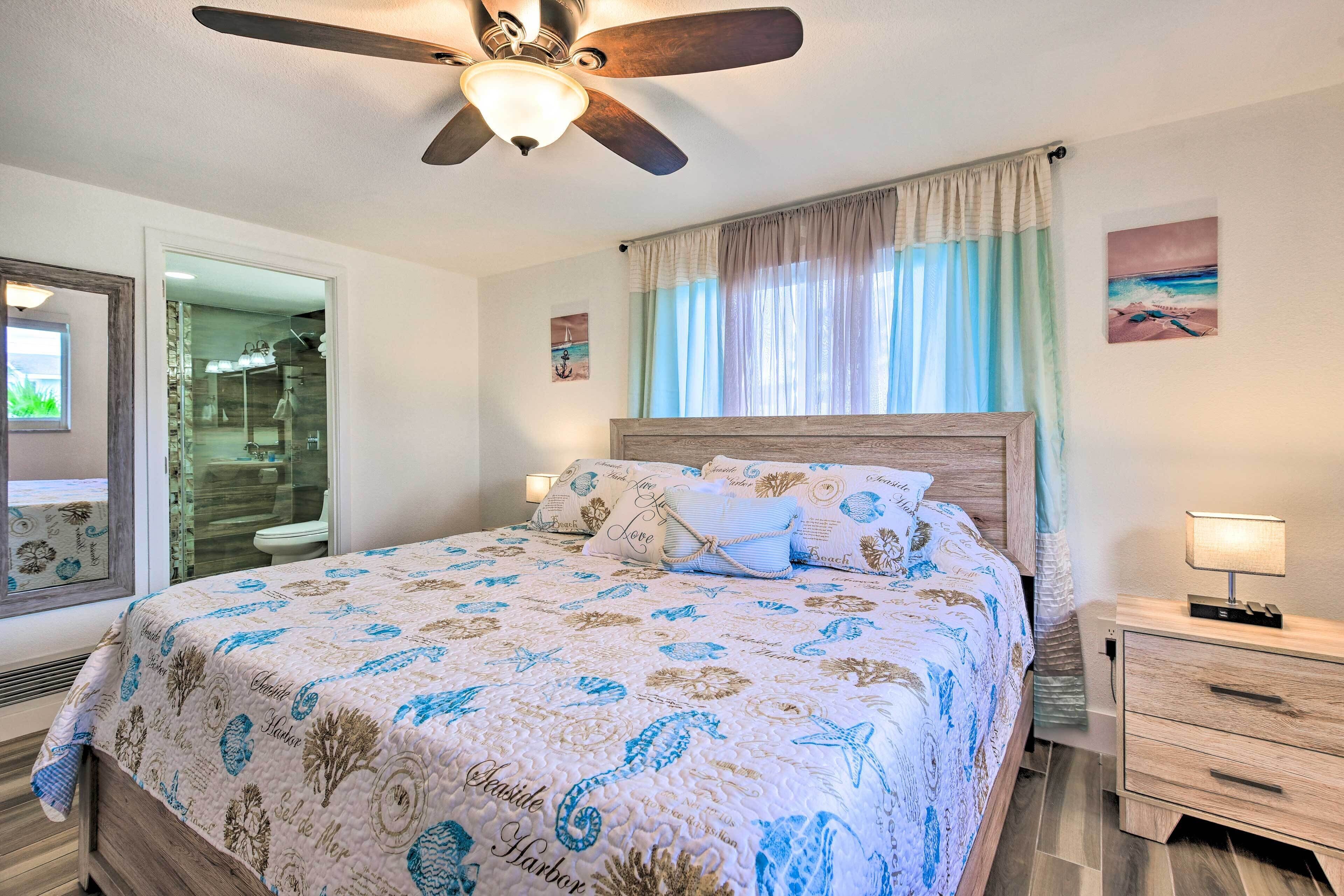 Un dormitorio con cama, mesita de noche, ventilador de techo, ventana con cortinas y un baño visible a través de una puerta abierta.