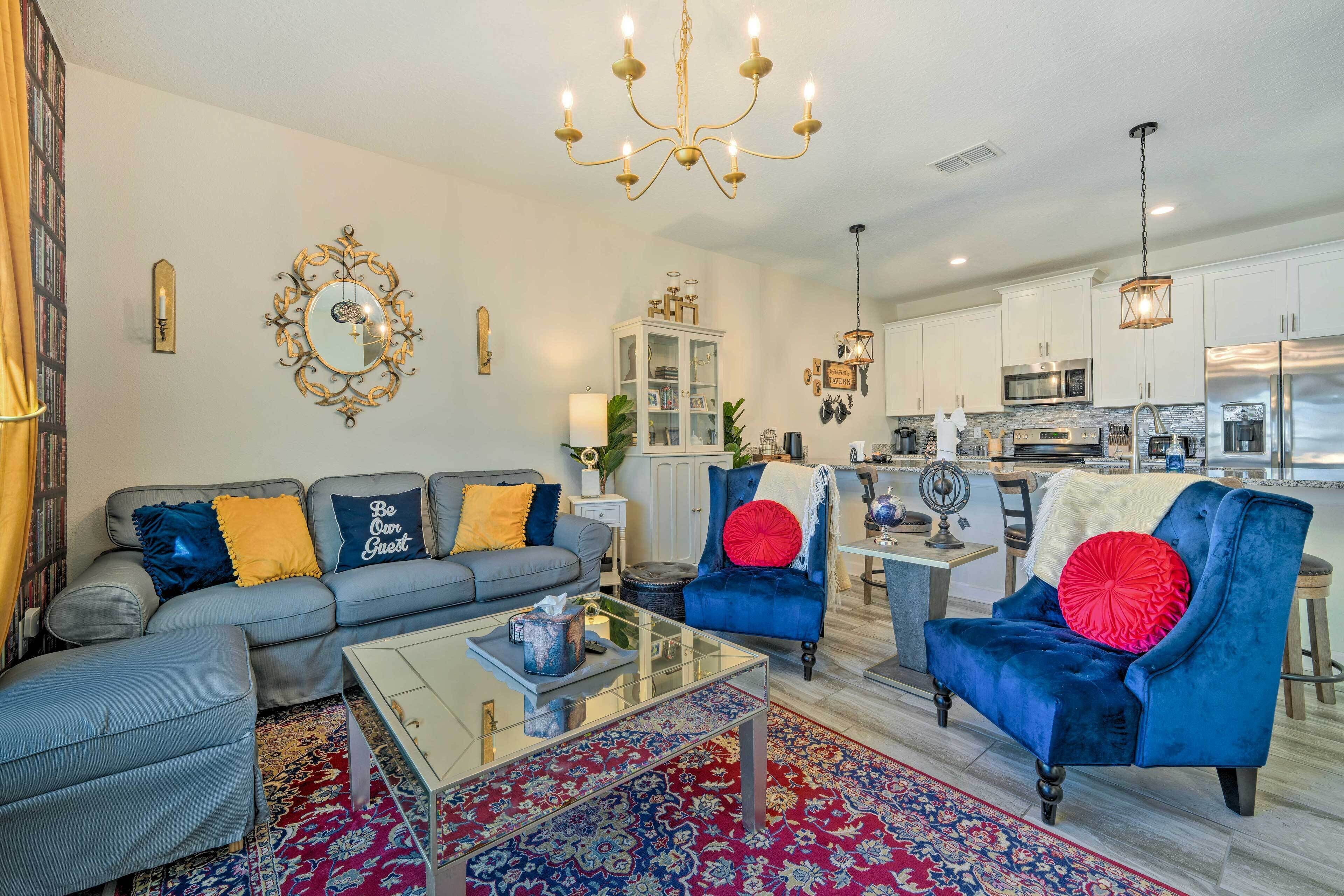 A modern living room with a grey sofa, a blue armchair, and a glass coffee table. There is a kitchen in the background with white cabinets and stainless steel appliances.