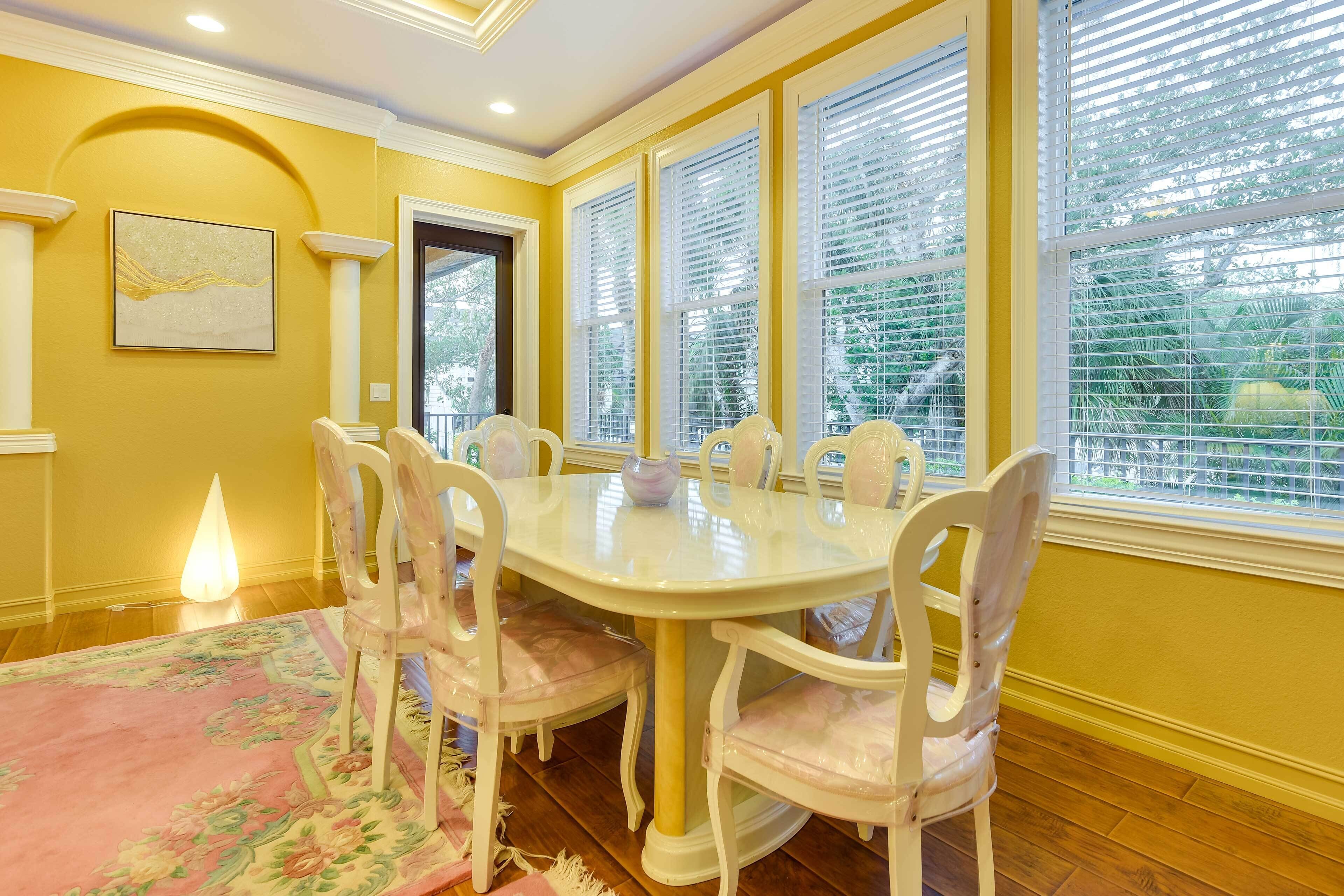 Une salle à manger avec une table ronde et six chaises, de grandes fenêtres équipées de stores, un tapis à motifs floraux et un luminaire fixé au mur.