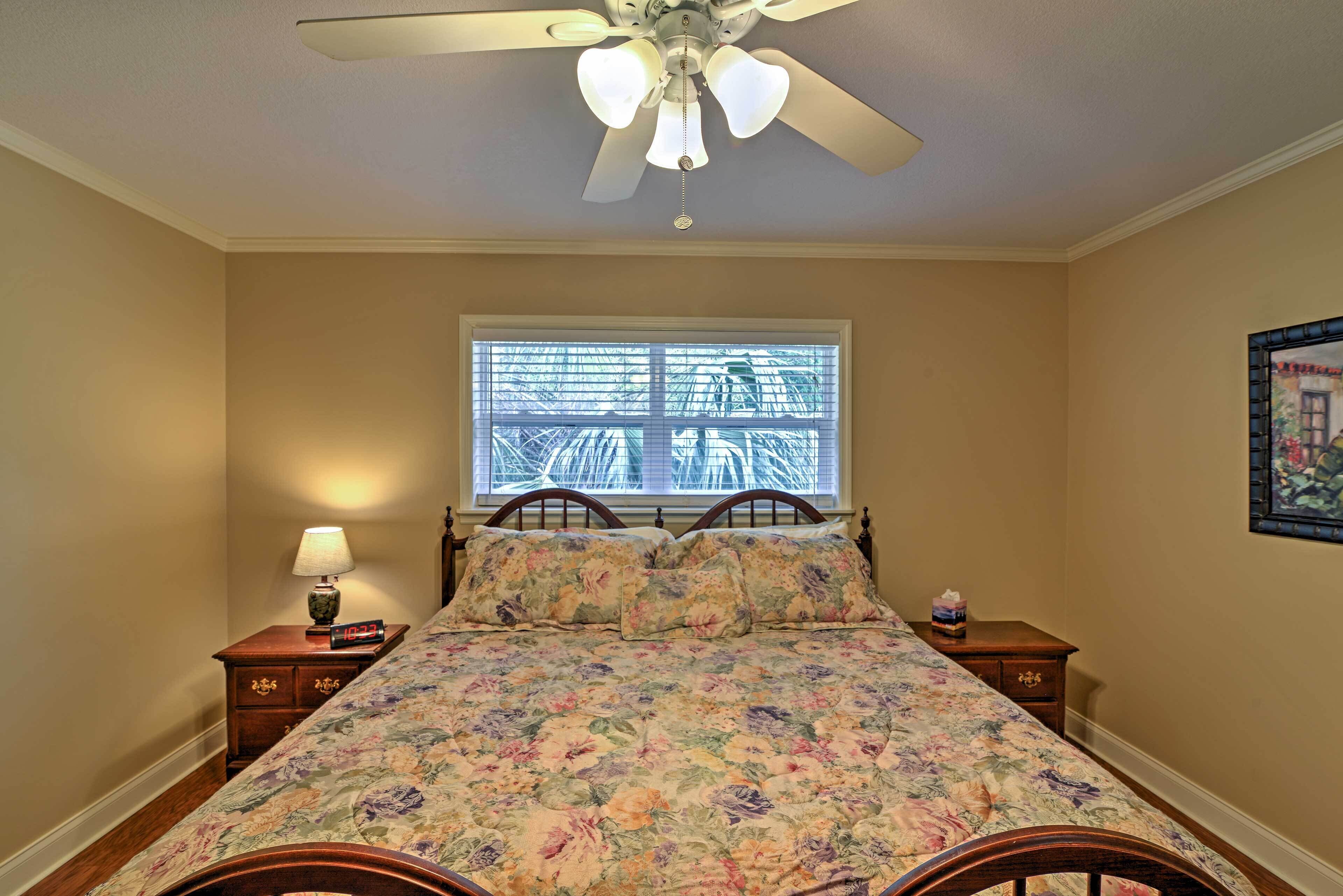 A bedroom with a bed, a ceiling fan, a nightstand with a lamp, a window with blinds, and a framed picture on the wall.
