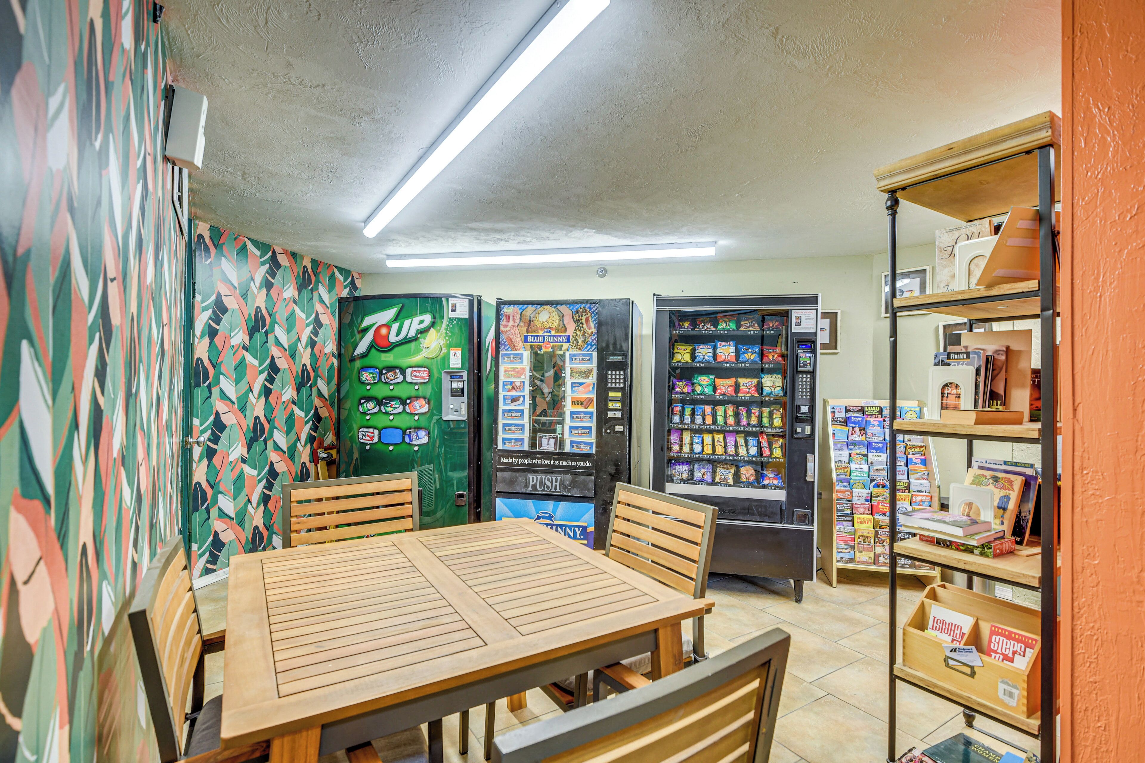 Una habitación con una mesa y sillas de madera, máquinas expendedoras y una estantería con libros y revistas.