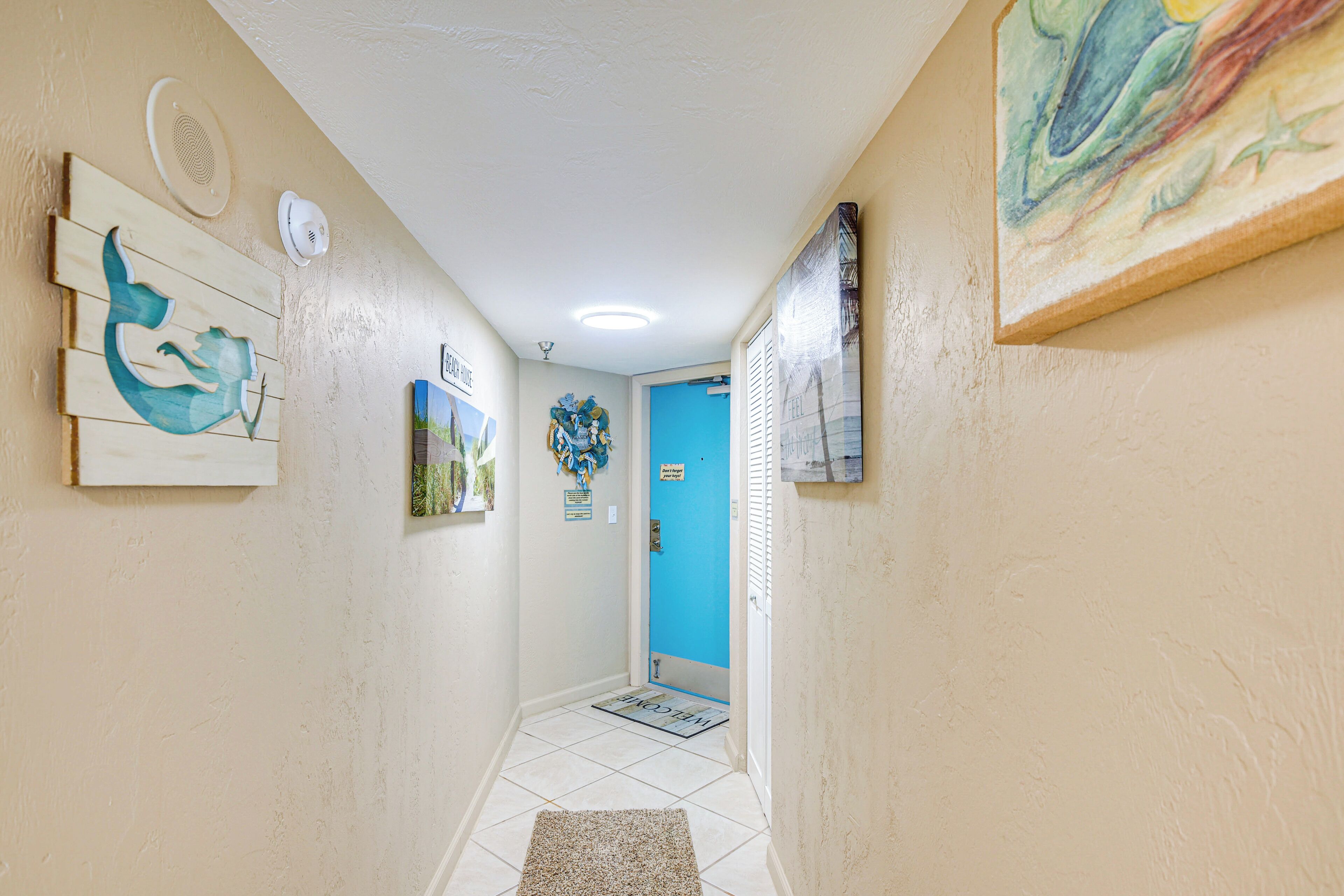 A hallway with a blue door, a welcome mat, and various wall decorations including a whale mural and a framed picture.