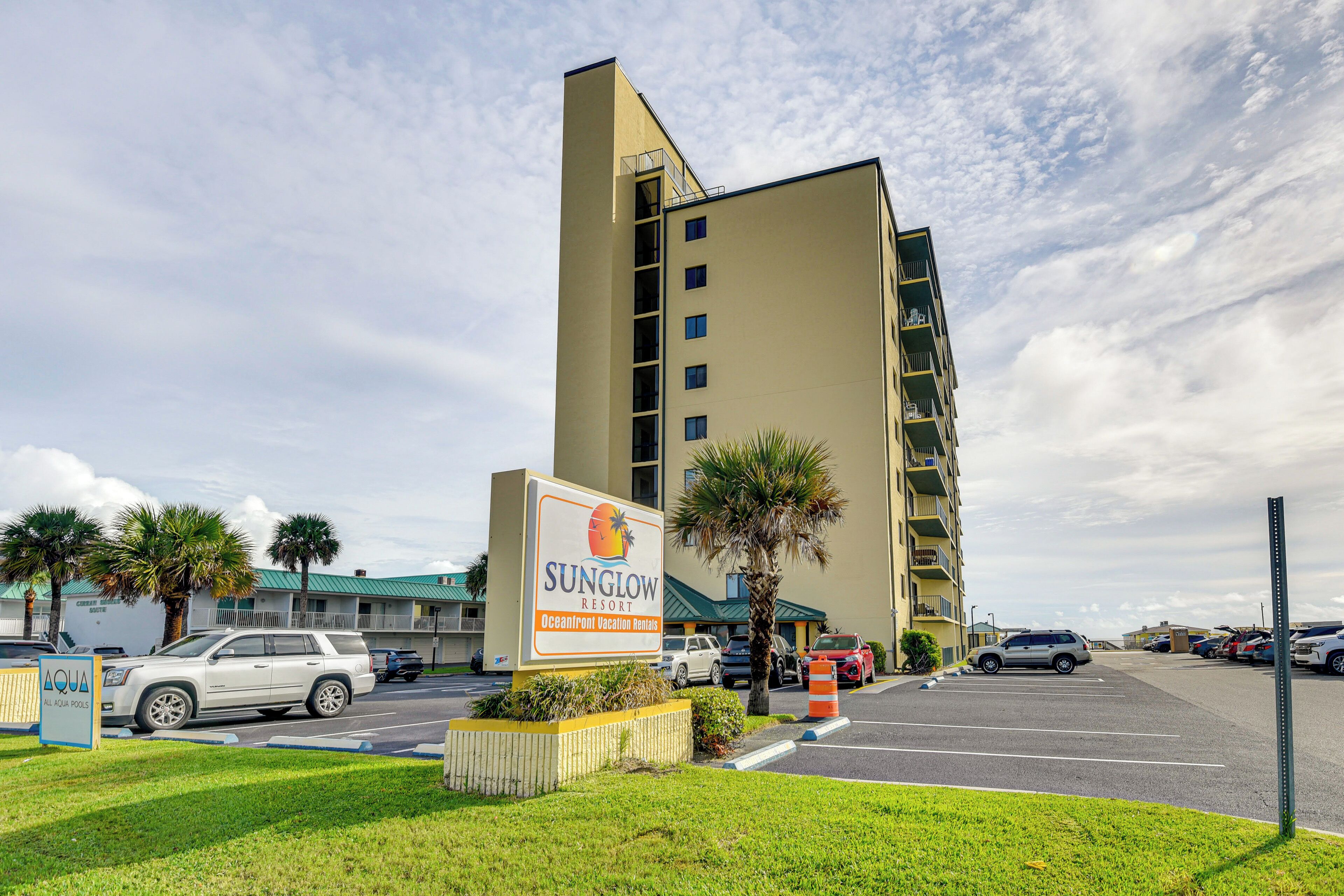 Un hôtel avec une enseigne indiquant « Sunglow Resort, location de vacances en bord de mer » et un parking où sont garées plusieurs voitures.