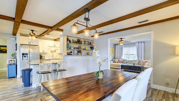 A spacious dining area with a wooden table, white chairs, and a ceiling fan. A kitchen with white cabinets and a refrigerator is visible in the background. - Florida Vacation Rental w/ Screened Lanai (Ocala)