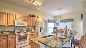 A kitchen with wooden cabinets, a granite countertop, a built-in oven, a microwave, a refrigerator, and a dining area with wicker chairs. - ~ 9 Mi to Disney: Family Home w/ Private Pool! (Davenport)