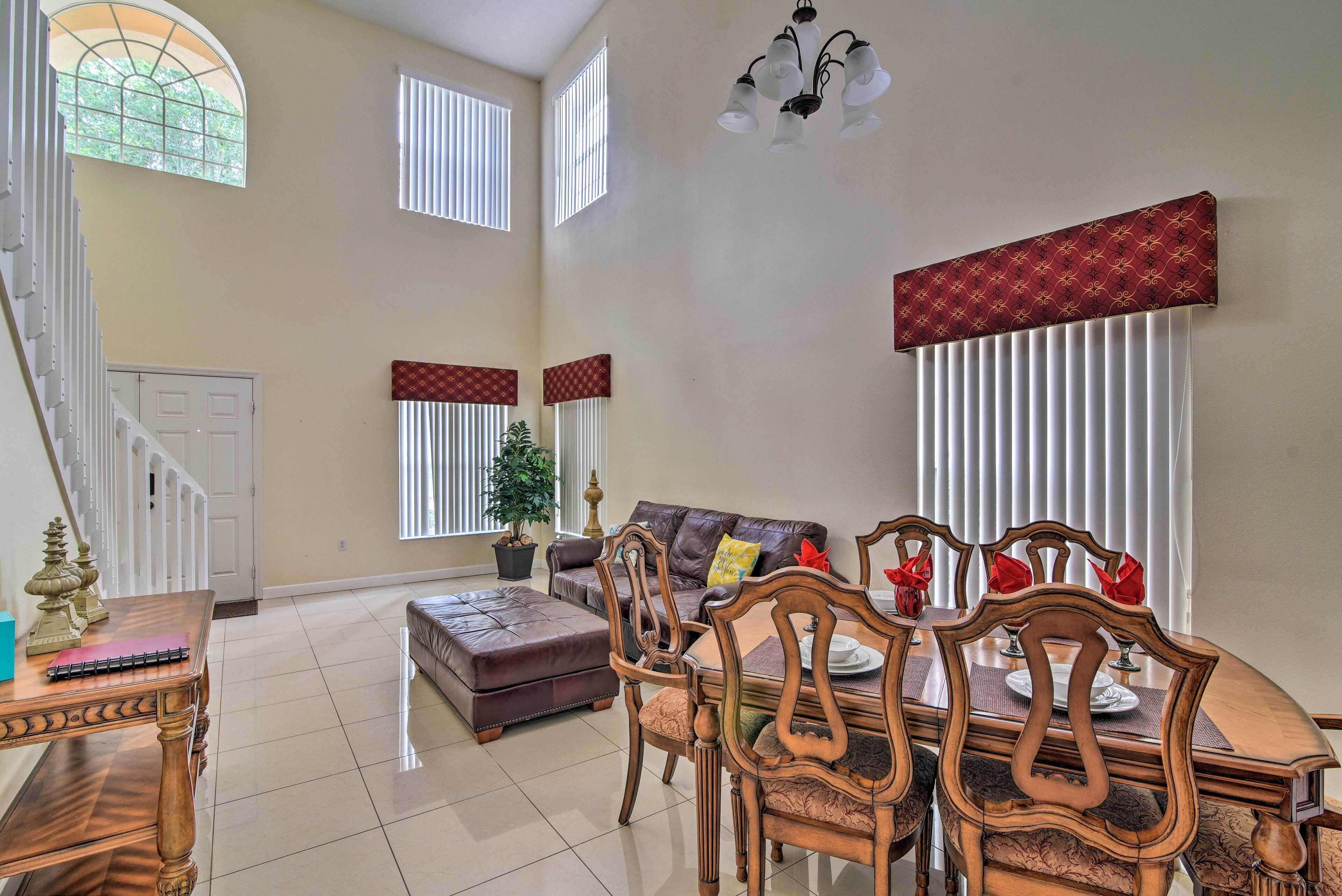 A spacious living room with a dining area, large windows, a chandelier, and a staircase.
