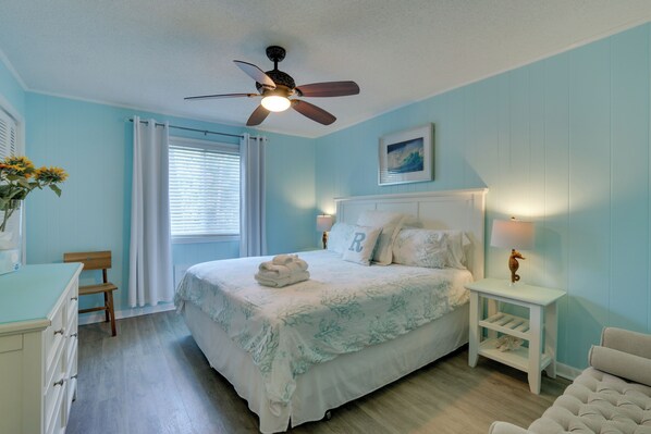 A bedroom with a bed, a nightstand, a chair, a desk, a ceiling fan, and a window with curtains.