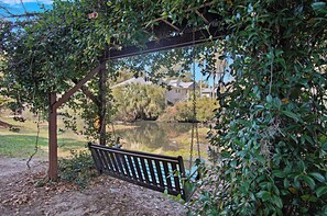 A wooden swing bench under a pergola with a view of a lake and houses in the background.