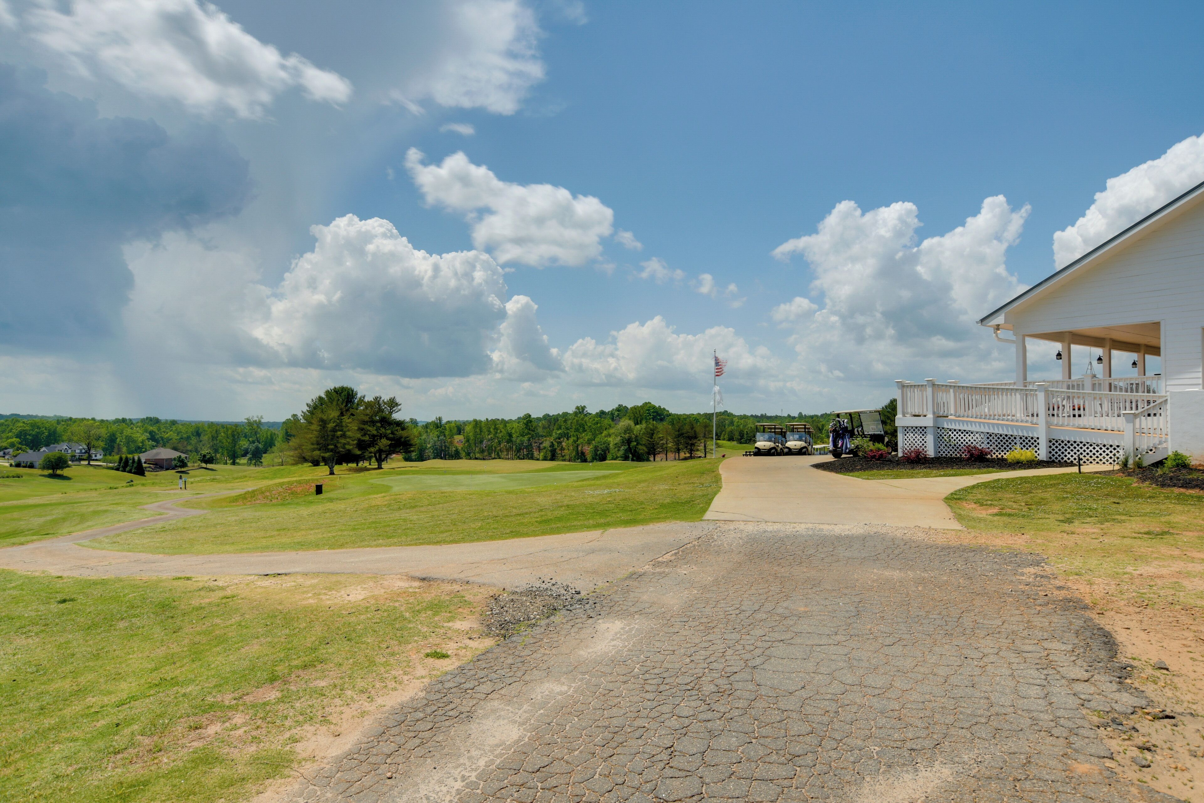 A paved driveway leading to a white building with a porch, surrounded by a grassy area and trees.