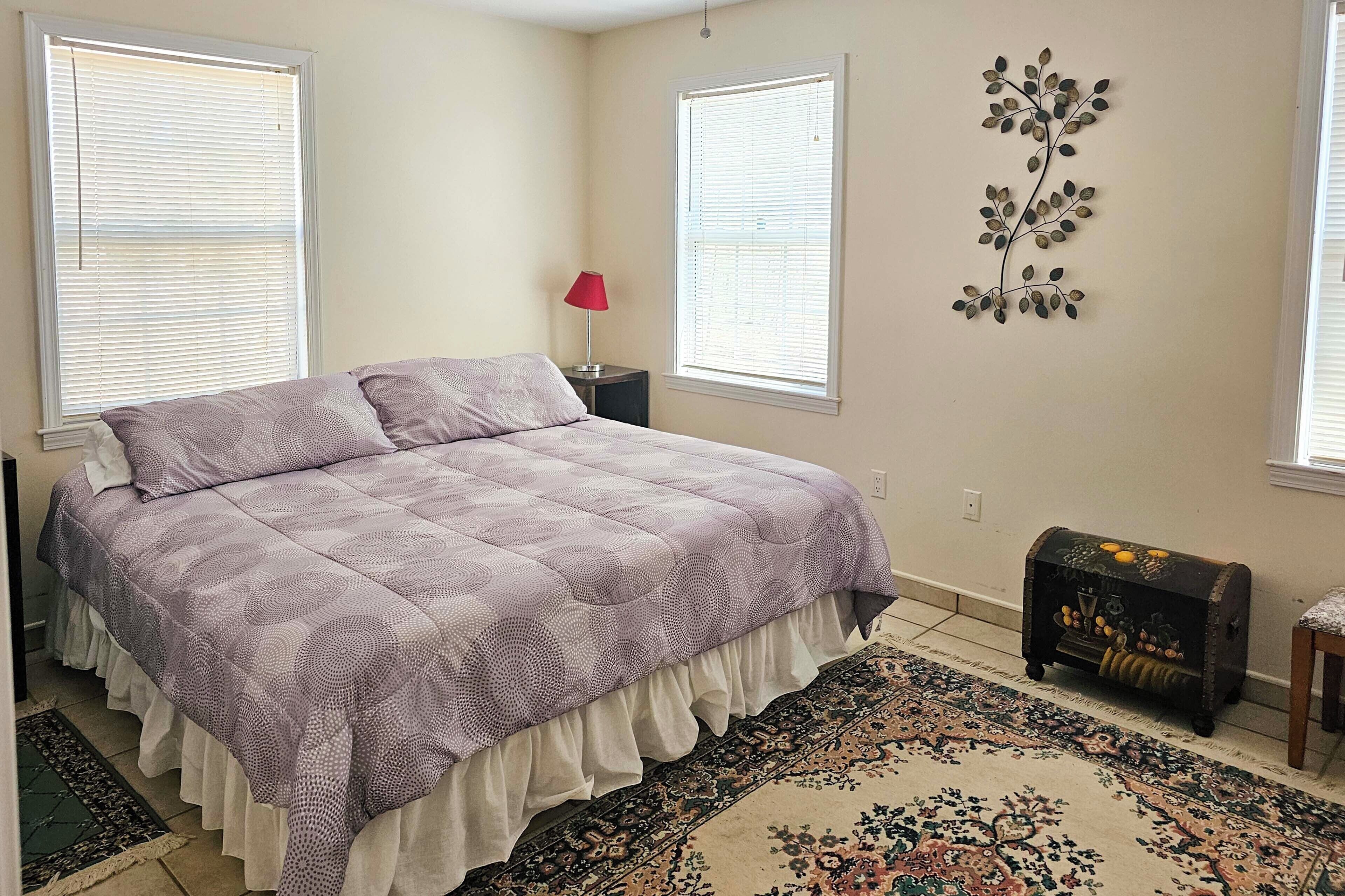 Une chambre à coucher comprenant un lit, deux fenêtres, une table de chevet, une lampe, un élément décoratif mural, un tapis et une petite commode.
