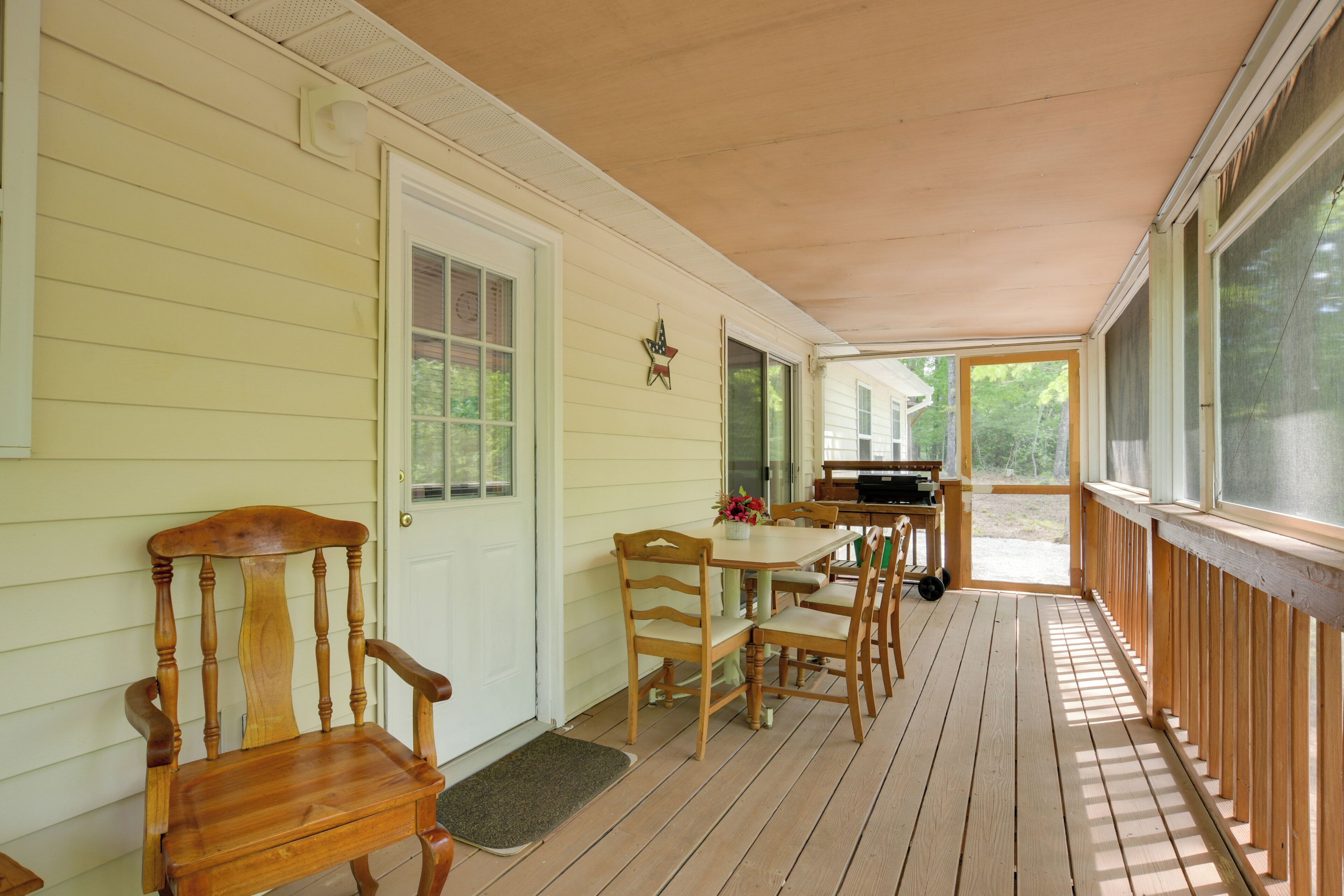 A covered porch with a wooden table and chairs, a door, and a window overlooking greenery.