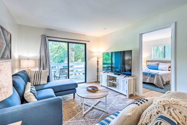 A modern living room with a sofa, coffee table, and TV. There is a balcony with a view of trees.