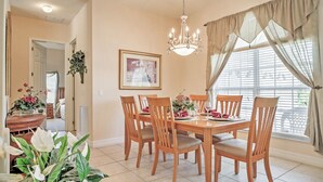 Une salle à manger avec une table en bois dressée pour six personnes, entourée de six chaises. Un lustre est suspendu au plafond et un tableau encadré orne le mur.