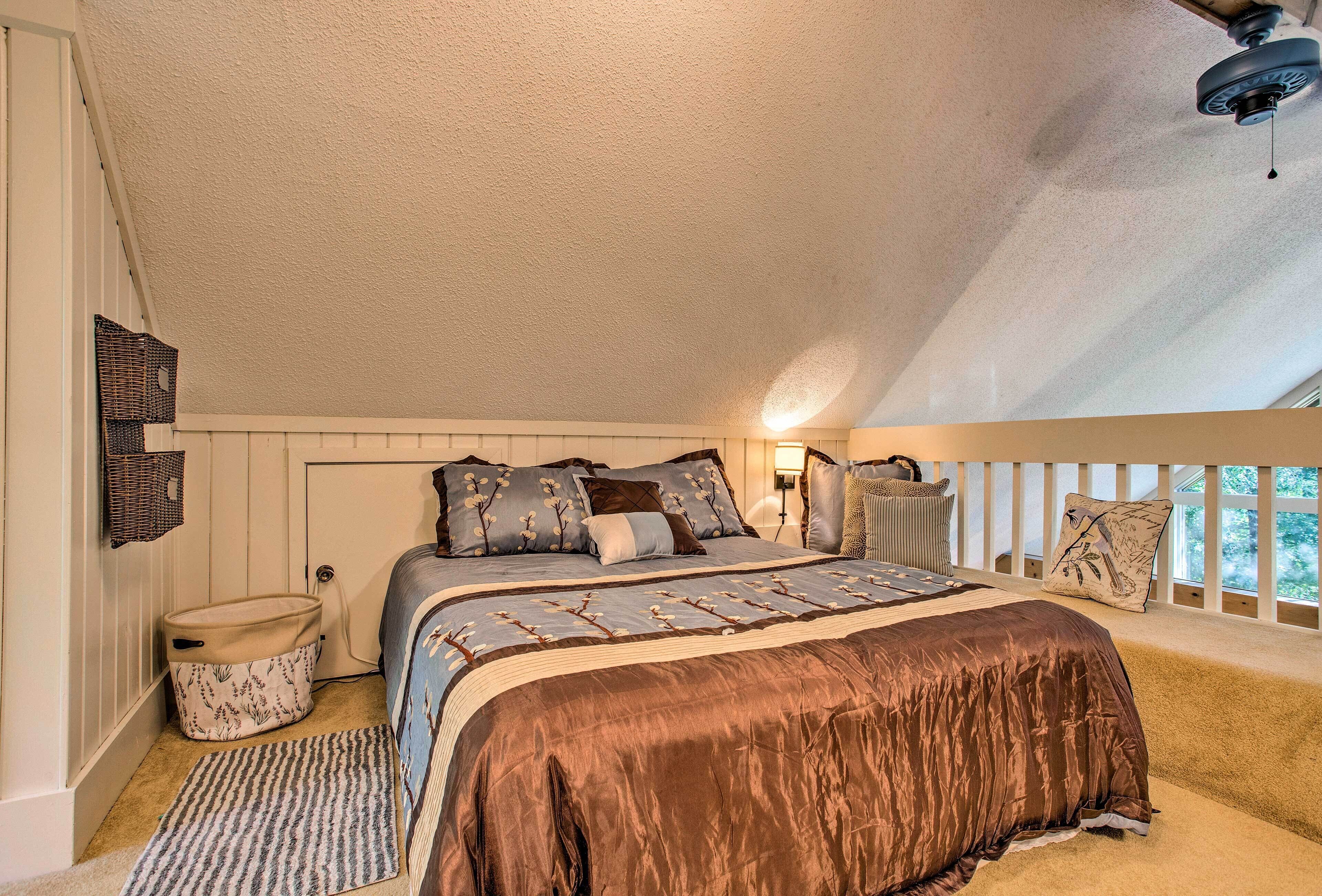 Une chambre avec un plafond en pente, un lit recouvert d’une couverture marron, une table de chevet, une lampe et une fenêtre donnant sur un beau paysage.
