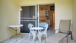 Uma pequena área de pátio com uma mesa e duas cadeiras, um sofá e uma porta de vidro deslizante que dá para um quarto.
