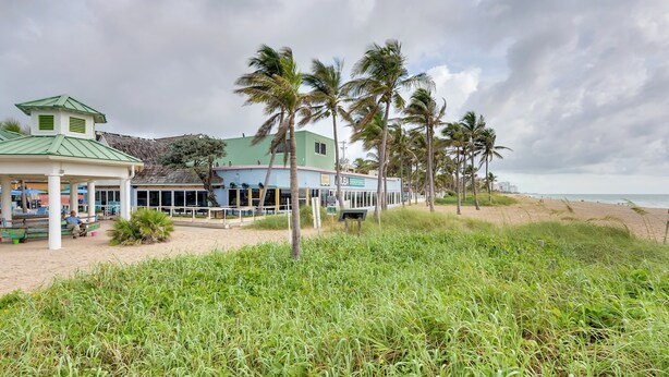 Um hotel à beira-mar com um pavilhão de telhado verde, palmeiras e uma praia de areia.