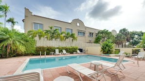Piscina com espreguiçadeiras e cerca branca, cercada por palmeiras e vegetação.
