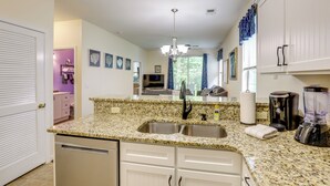 A kitchen with granite countertops, a double sink, white cabinets, a chandelier, and a view of the living room.