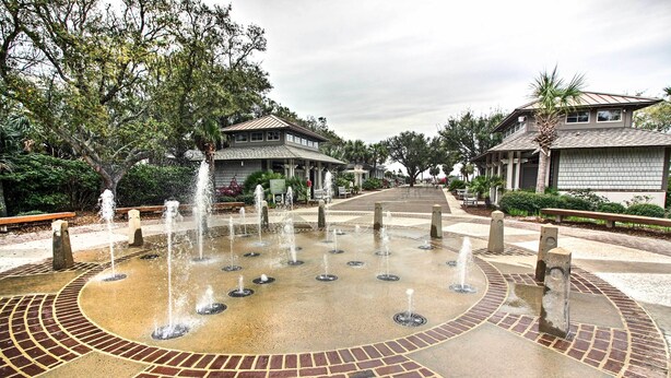 A circular water fountain with multiple jets, surrounded by a brick pathway and greenery.