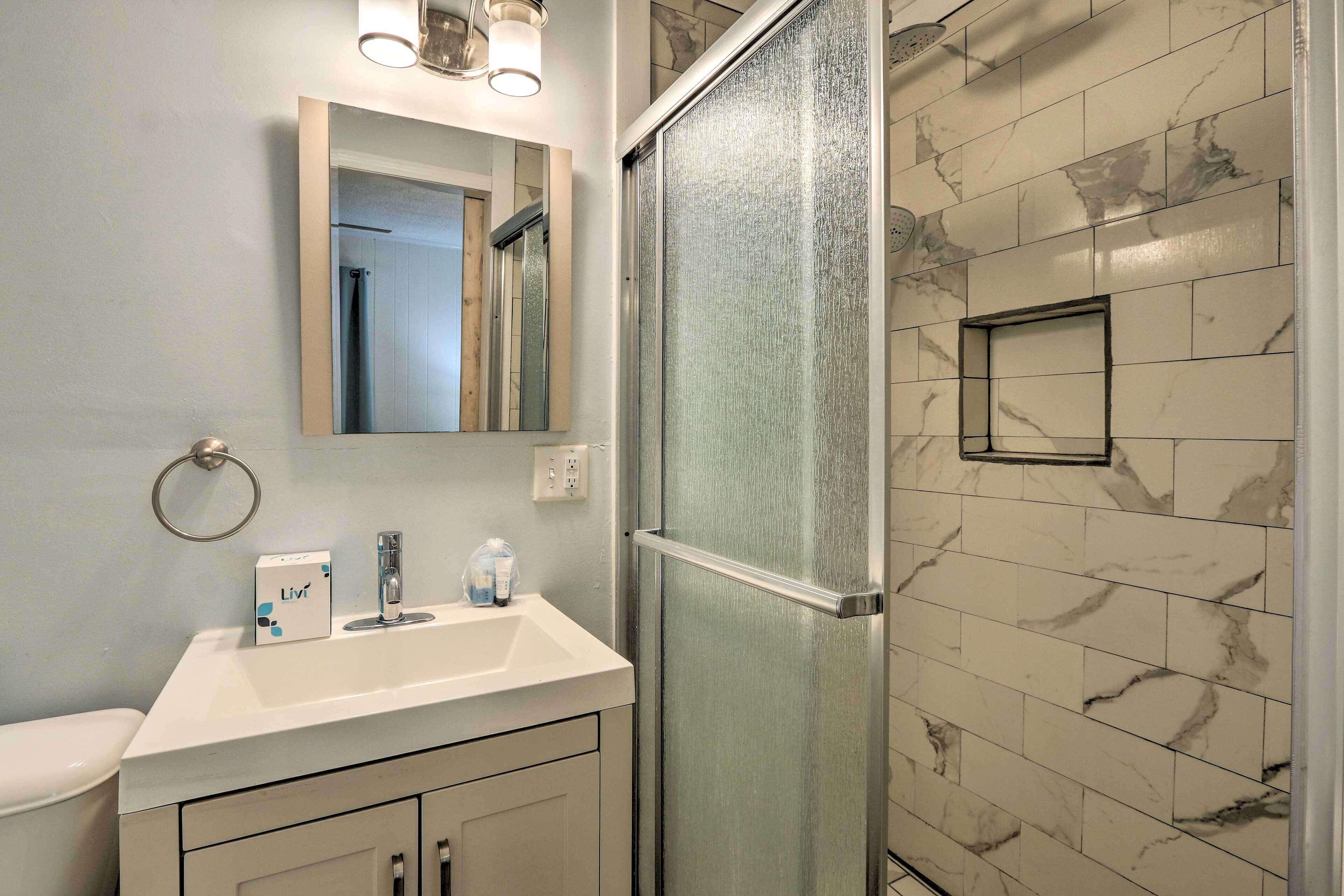A bathroom with a glass shower enclosure, a white sink with a mirror above it, and a marble-tiled wall.