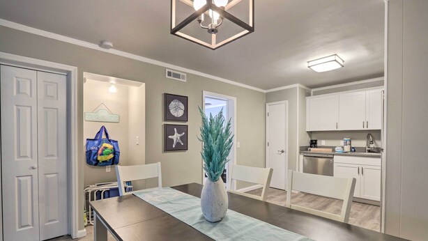 Un comedor con una mesa larga, sillas, un jarrón con planta y una pared decorada con cuadros y un letrero que dice 'BEACH PLEASE'.