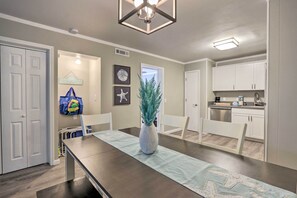 A dining area with a long table, chairs, a vase with a plant, and a wall with framed pictures and a 'BEACH PLEASE' sign.