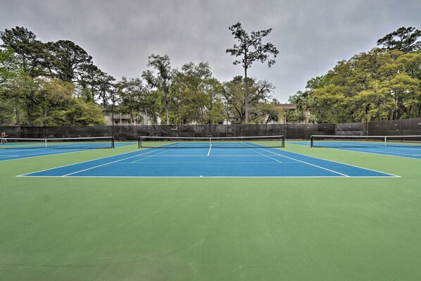 Una cancha de tenis exterior con varias canchas, rodeada de árboles y una valla.