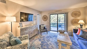 A living room with a sofa, armchair, and coffee table, a television, and a large window with a view of trees. - Half-mi to Beach: Hilton Head Condo w/ Lagoon View (Hilton Head Island)