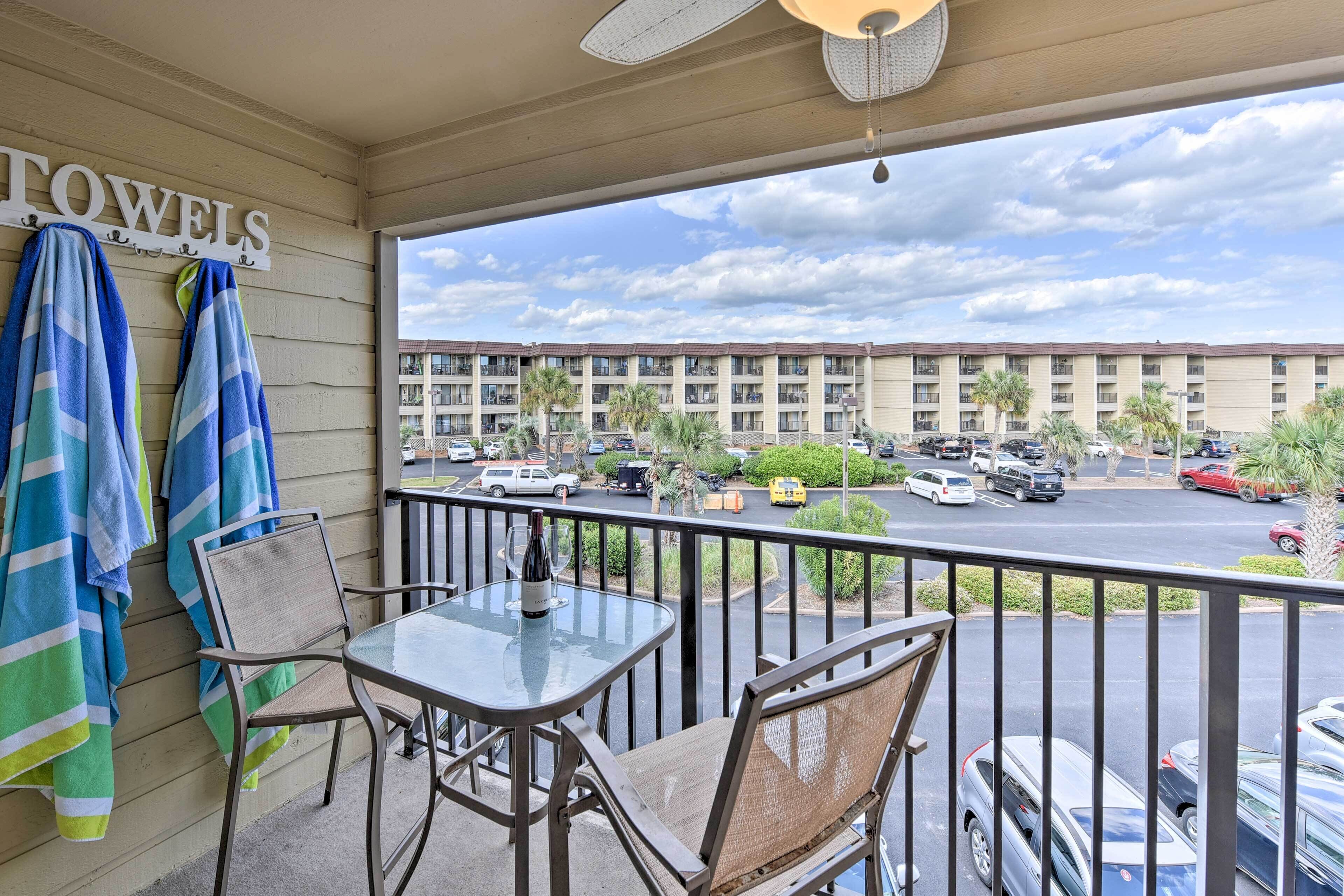 A balcony with a table and chairs, a sign reading "TOWELS", and a view of a parking lot and buildings in the background.