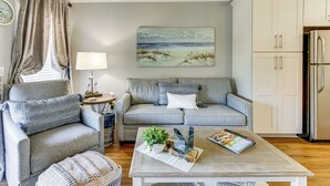 Een woonkamer met een grijze bank, een salontafel met tijdschriften en een plant, een schilderij van een strandgezicht, en een keuken met een koelkast.