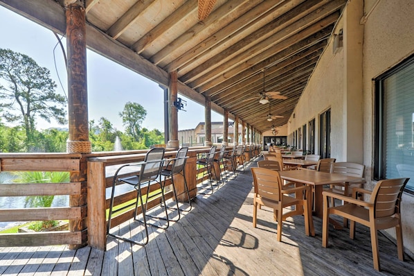 A covered outdoor seating area with wooden tables and chairs, overlooking a body of water and greenery.
