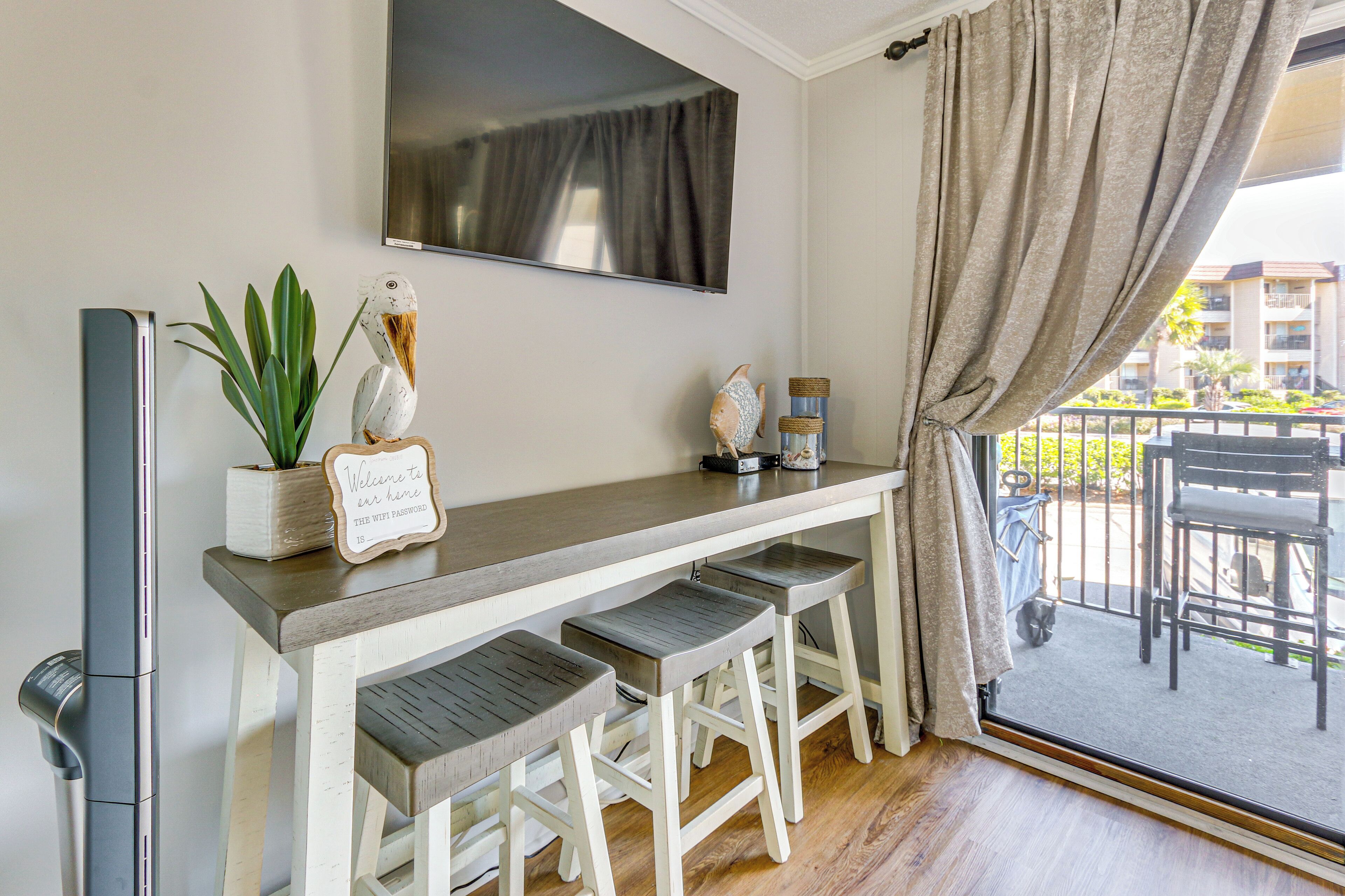 A room with a flat-screen TV mounted on the wall, a small table with a potted plant, a sign with a message, and a view of the outside balcony.