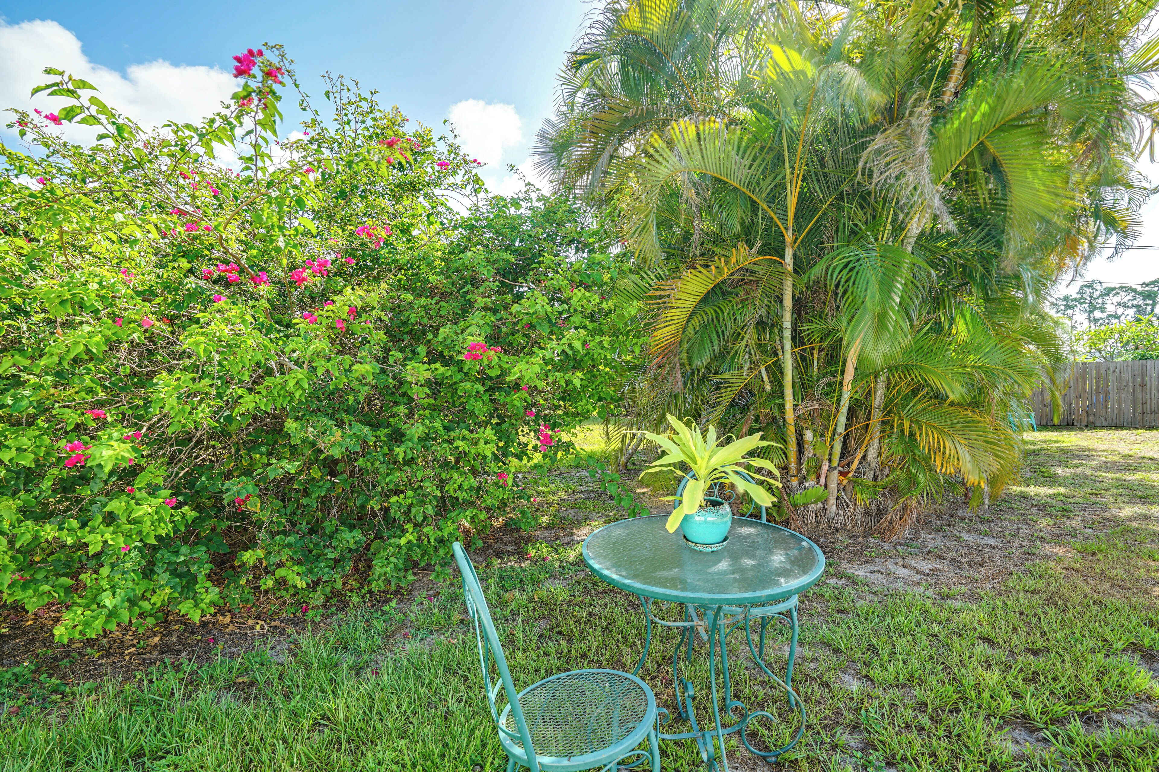 A small outdoor seating area with a table and chair, surrounded by lush greenery and a palm tree.