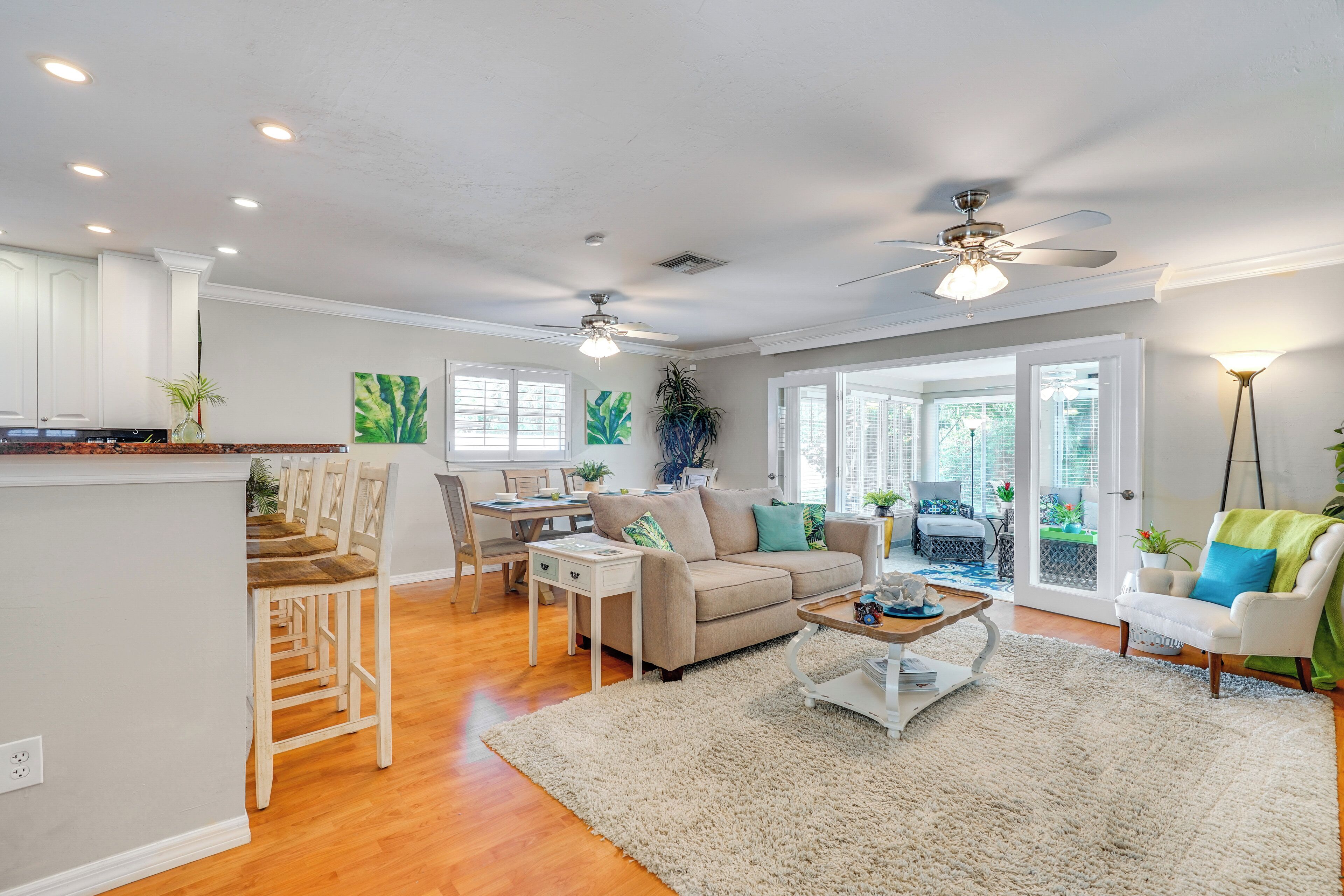 A spacious living room with a sofa, coffee table, and chairs. There is a kitchen area with a dining table and chairs. The room has large windows and a ceiling fan.