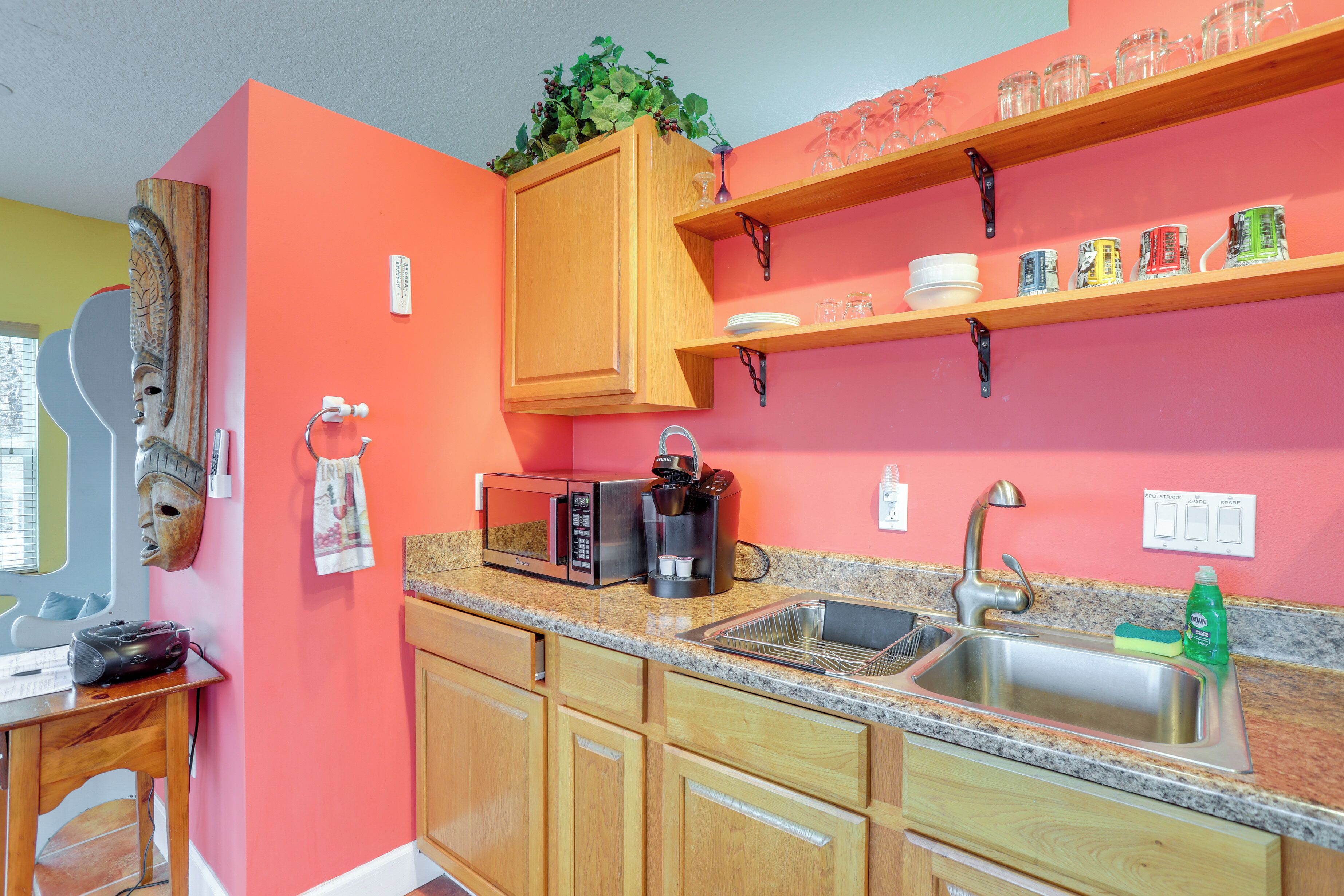 A kitchen with wooden cabinets, a granite countertop, a sink, a microwave, a coffee maker, and shelves with glassware.