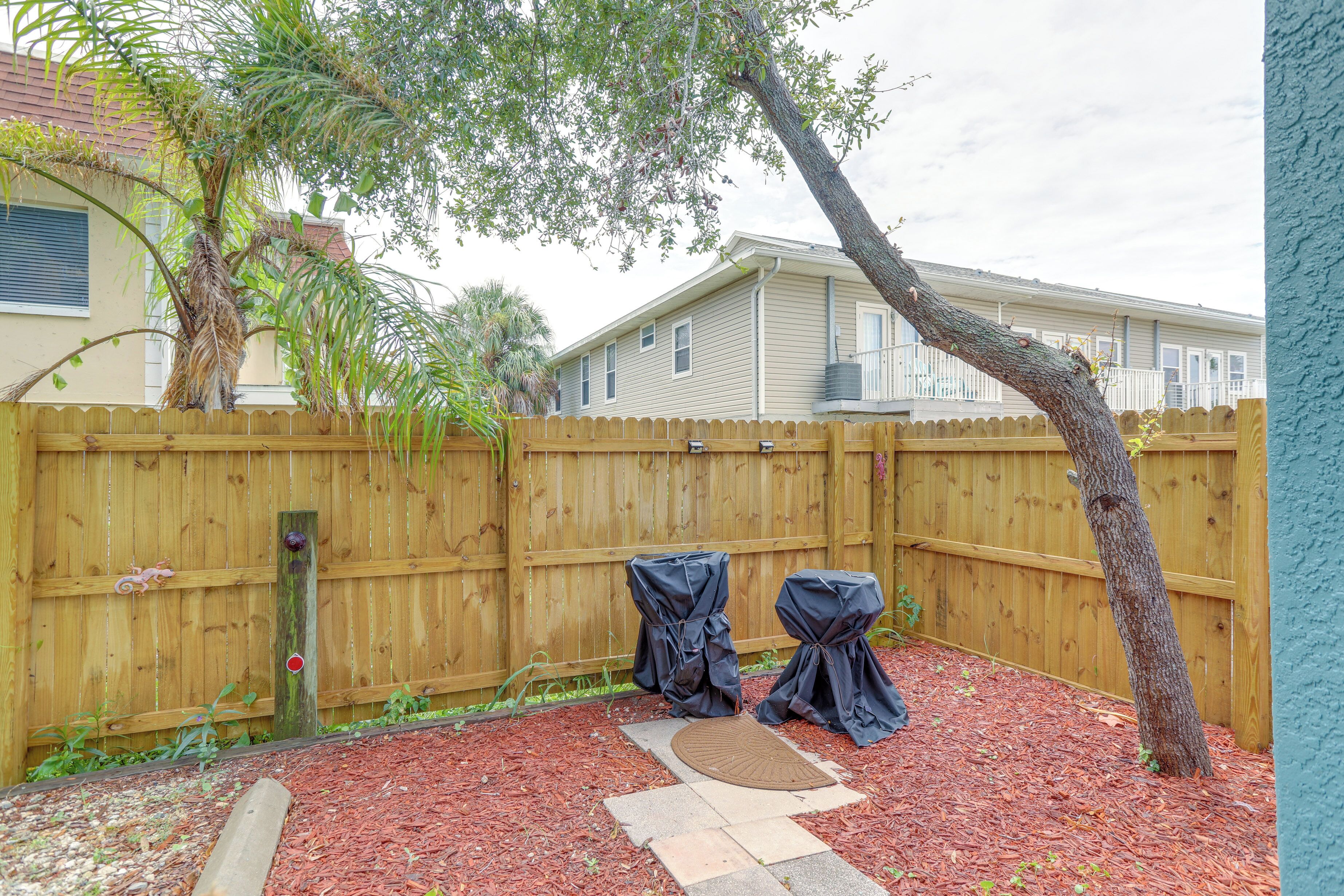 A backyard with a wooden fence, two covered barbecue grills, a palm tree, and a house in the background.