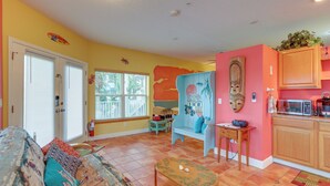 A living room with a sofa, a coffee table, and a small table with a chair. There is a window with blinds, a door, and a wall with a mural. - Vibrant Condo - Walk to Indian Rocks Beach! (Indian Rocks Beach)