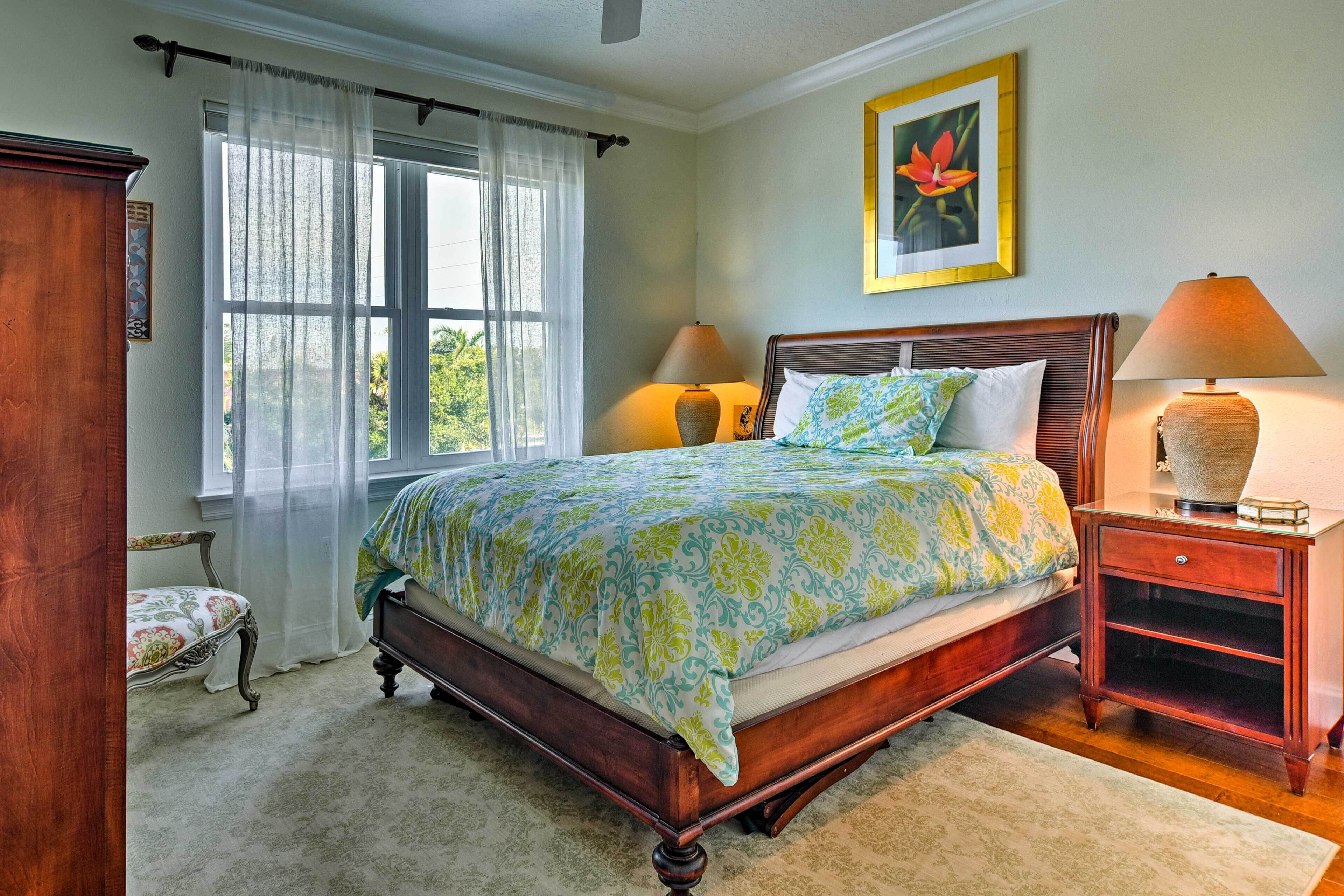 Une chambre avec un lit en bois, deux tables de chevet avec des lampes, une chaise, une fenêtre avec des rideaux légers et un tableau encadré au mur.