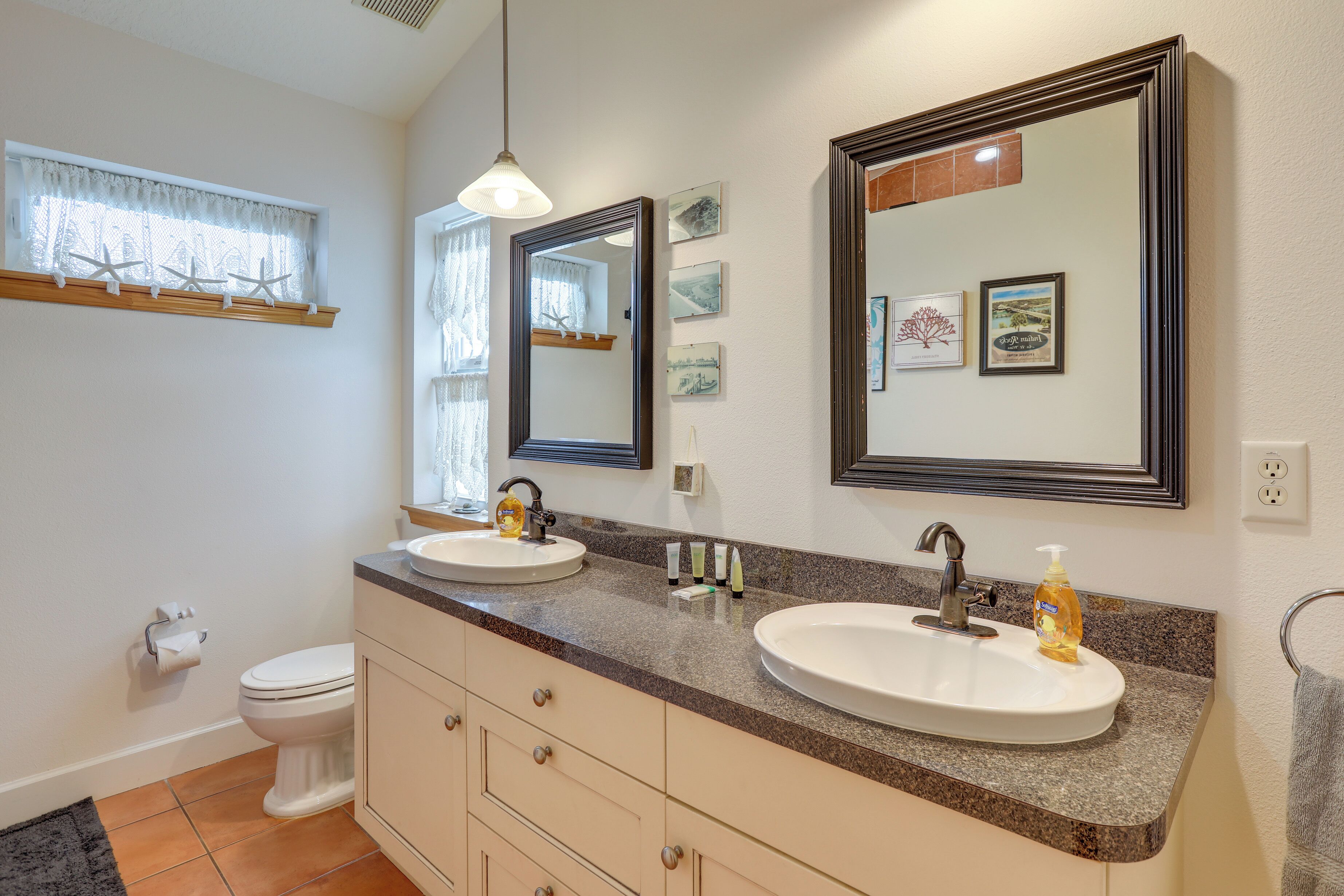 Une salle de bain avec deux lavabos, des toilettes, un meuble-lavabo avec des tiroirs, un miroir et une fenêtre décorée d’étoiles de mer.