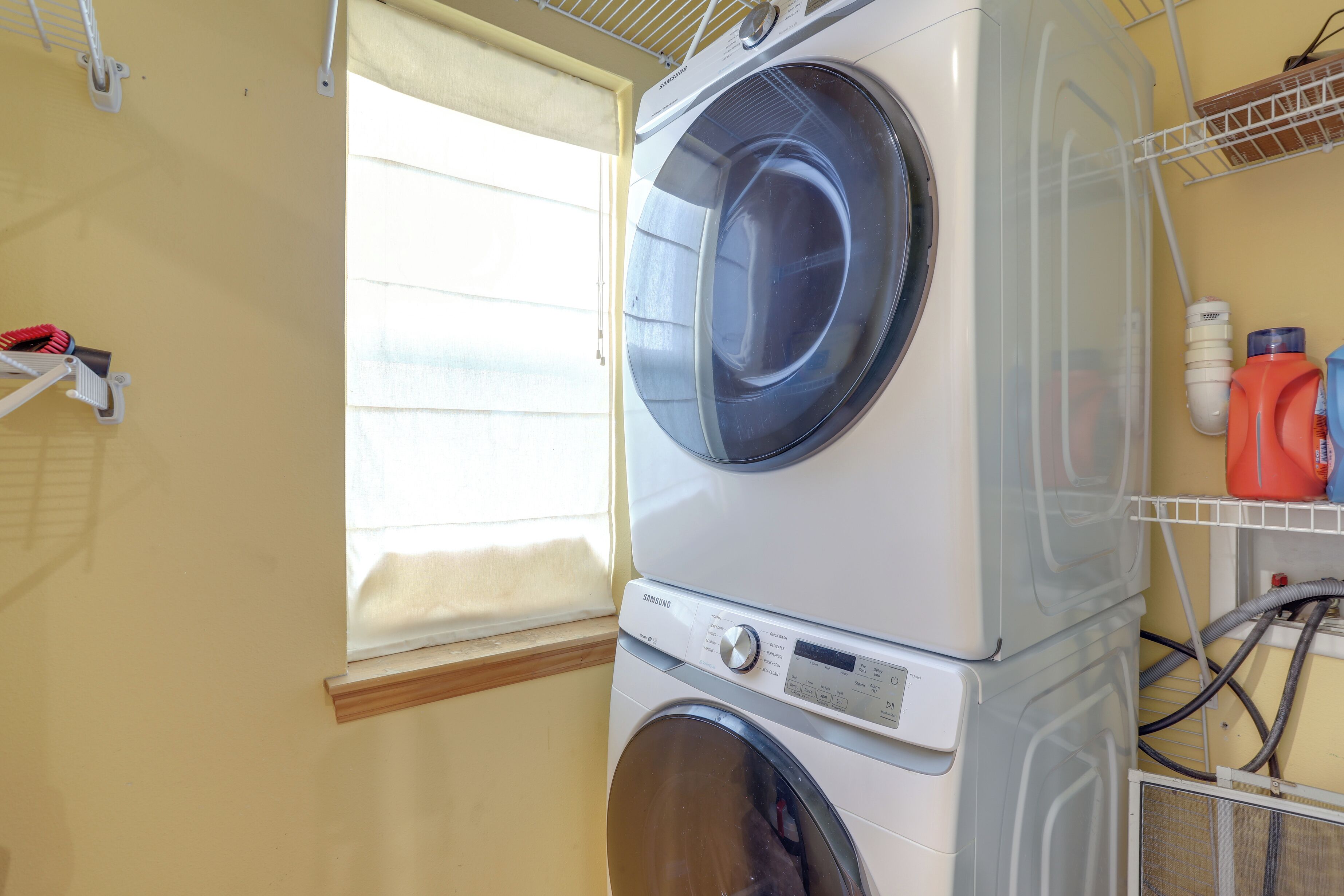 A stacked washer and dryer unit in a laundry room with a window and shelves holding cleaning supplies.