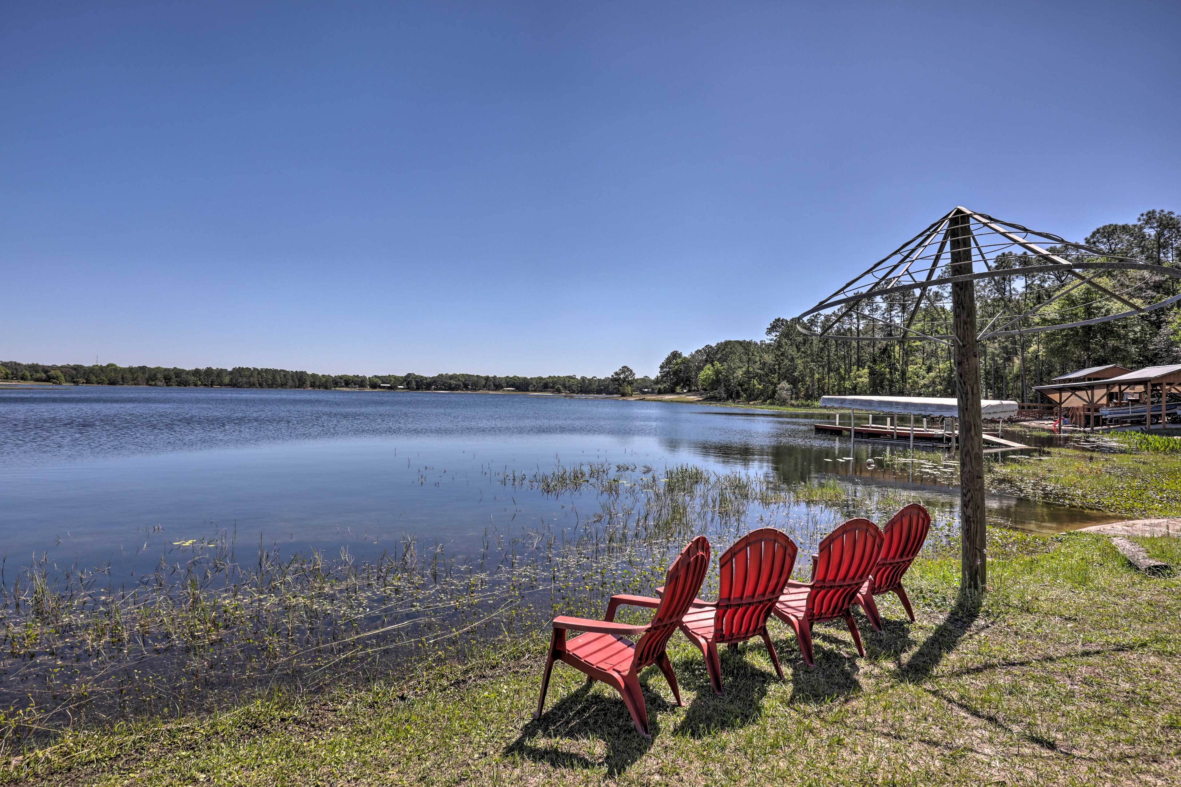 Blick auf einen See mit drei roten Adirondack-Stühlen, die dem Wasser zugewandt sind, einem Steg und einer Holzhütte mit Dach.