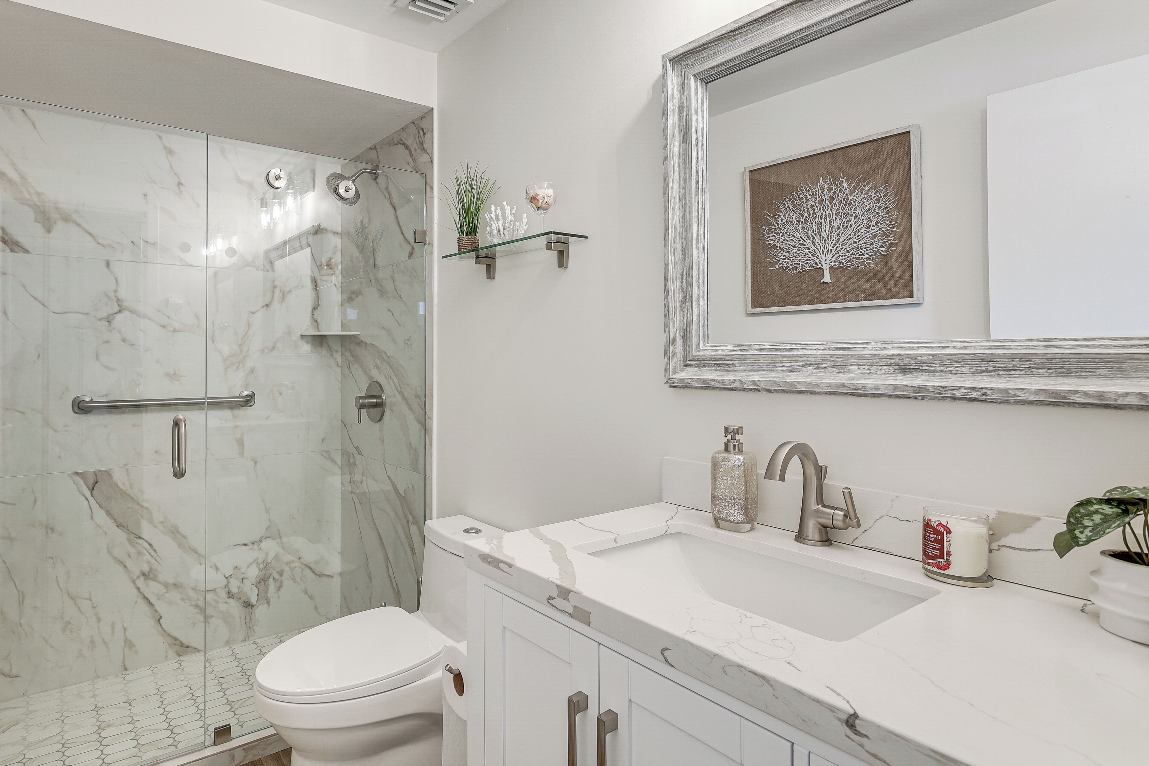 Une salle de bain dotée d’une douche en verre, de toilettes blanches, d’un lavabo recouvert de carreaux de marbre et d’un miroir orné d’une œuvre d’art encadrée.
