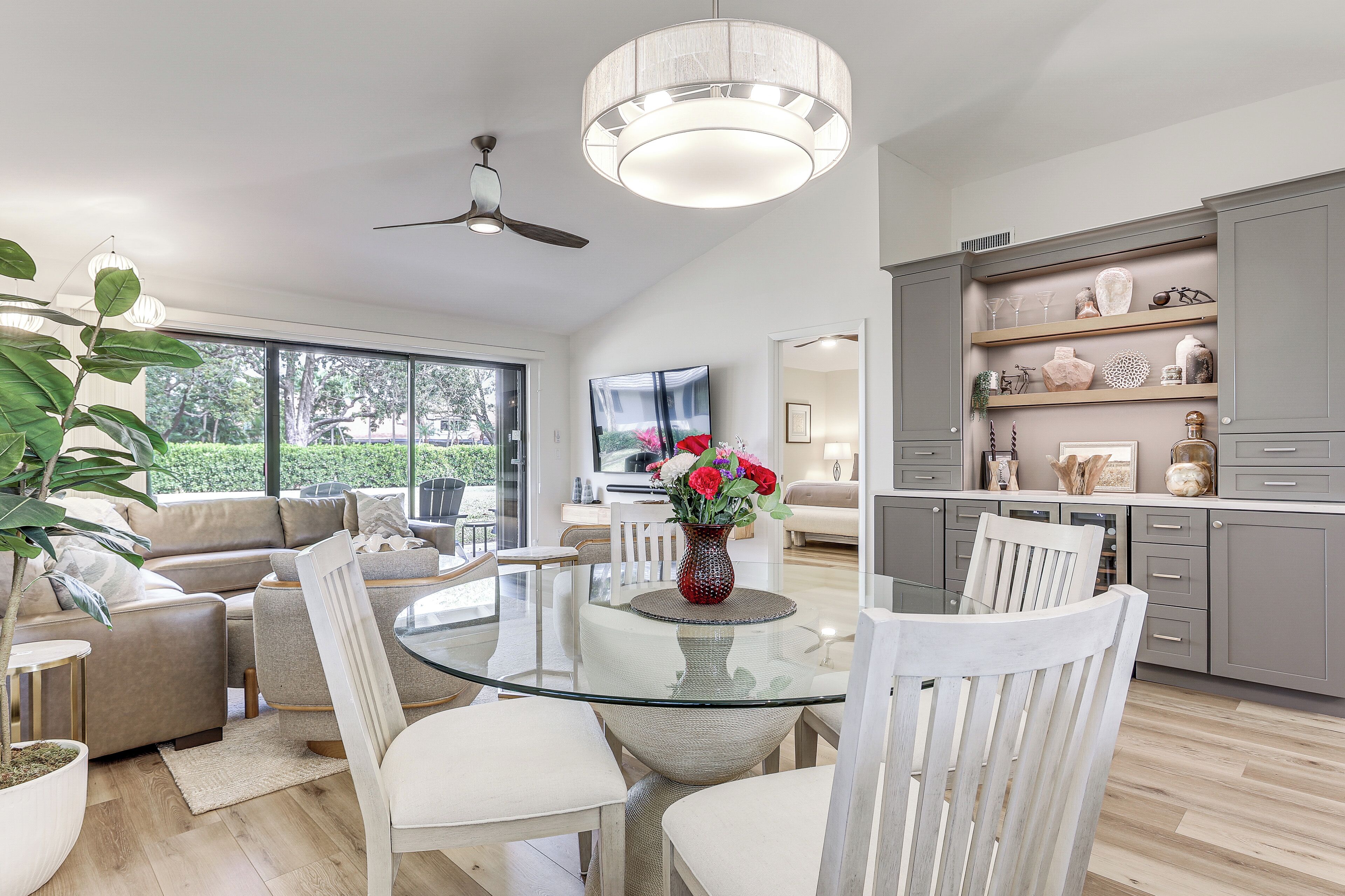 A modern living room with a glass dining table, grey cabinets, and a large window offering a view of the outdoors.
