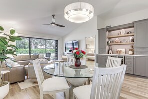 A modern living room with a glass dining table, grey cabinets, and a large window offering a view of the outdoors.